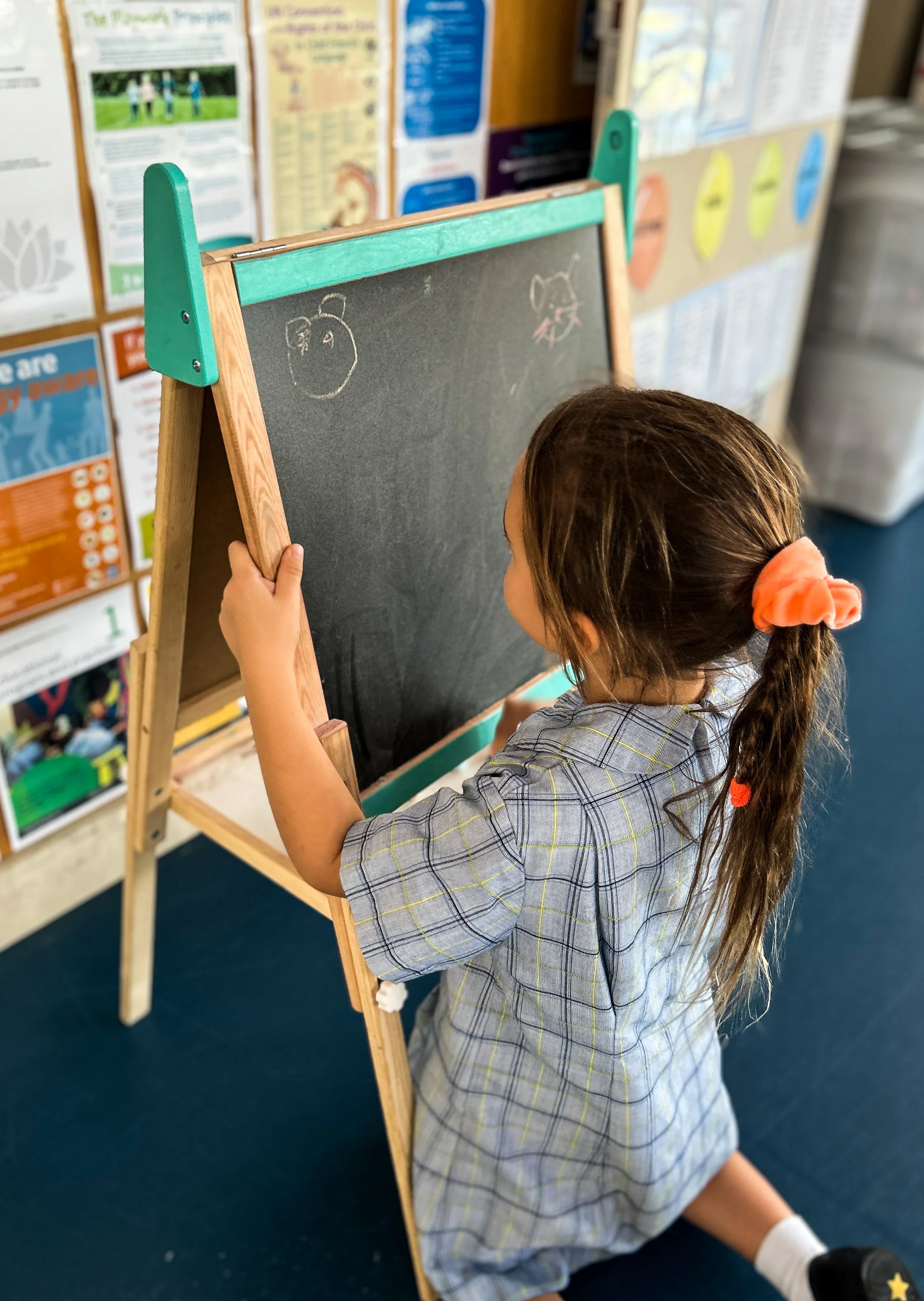 A young girl kneeling on a blue carpeted floor, drawing on a small chalkboard with chalk. She has long brown hair tied with an orange scrunchie and is wearing a school uniform. In the background, there are colorful educational posters on a bulletin b