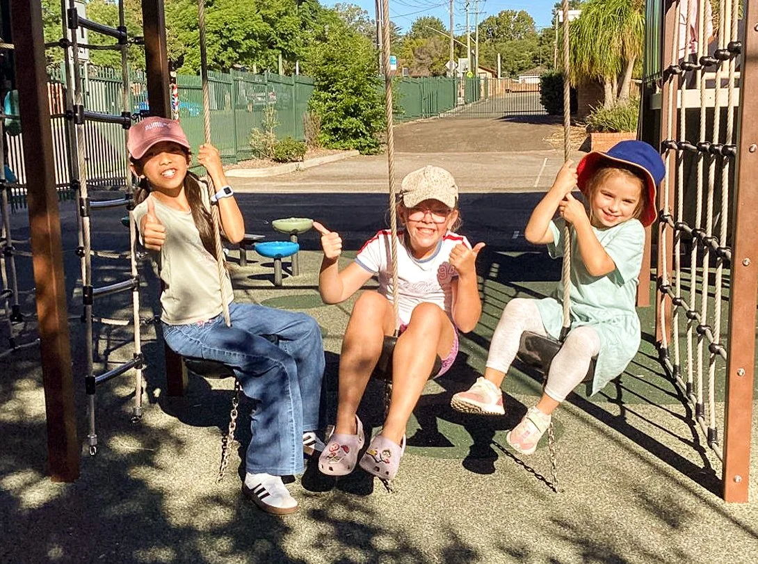 Three young girls smiling and playing on a playground swing set outdoors on a sunny day.
