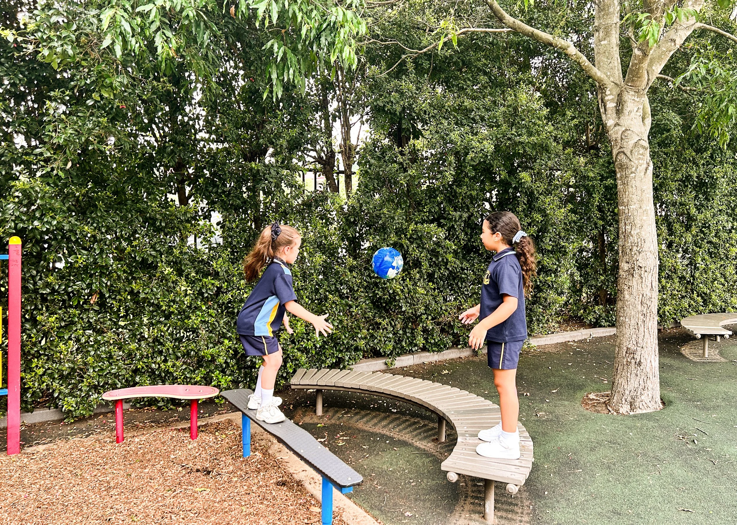 Two young girls in navy blue school uniforms playing on a curving wooden and metal balance beam in a lush green outdoor park with large trees and bushes.