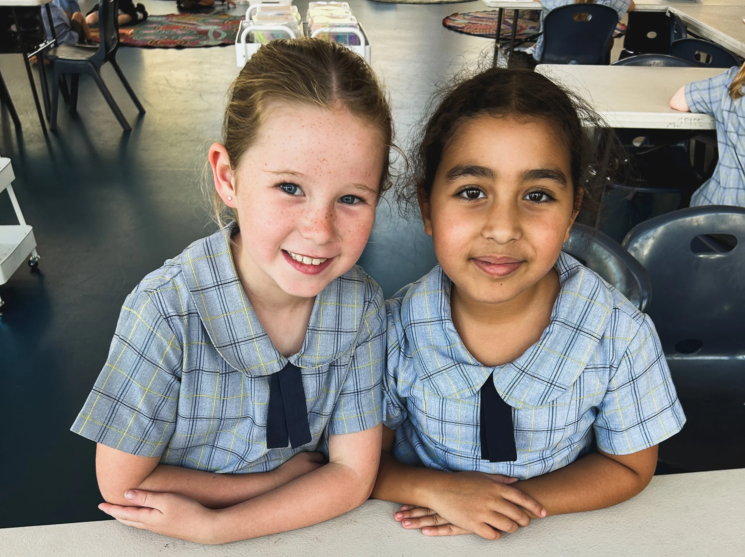 Two young girls wearing school uniforms sitting at a desk in a classroom, smiling at the camera.