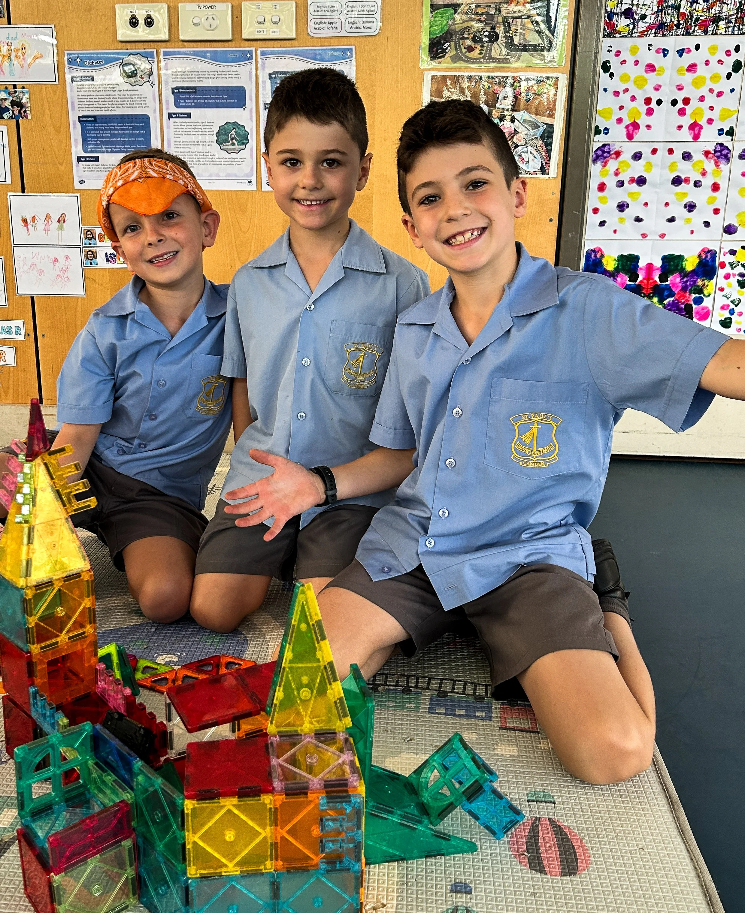Three young boys wearing blue school uniforms sitting on the floor in front of a colorful structure made from magnetic building tiles. Behind them is a bulletin board with educational posters and children's drawings.