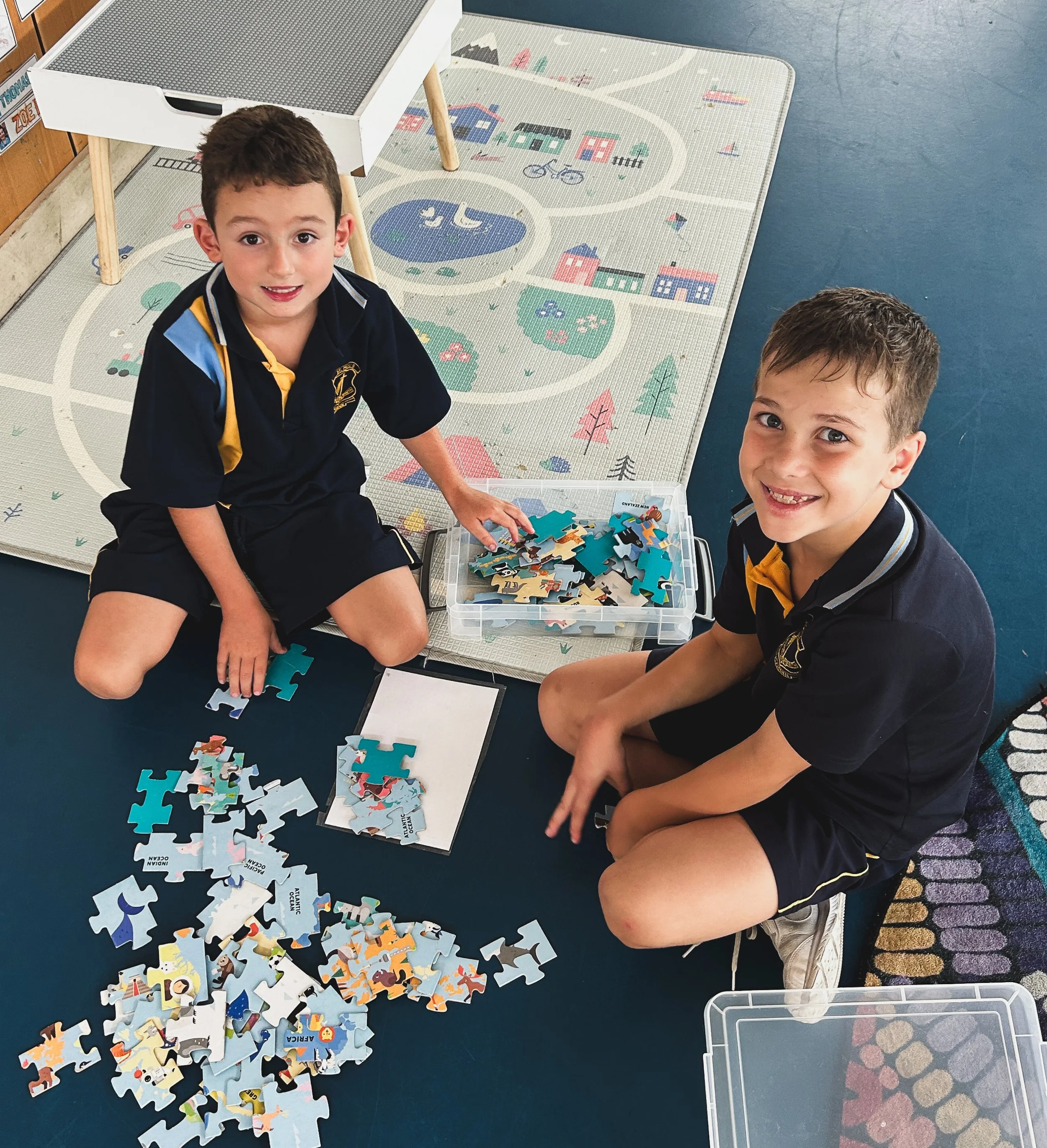 Two young boys sitting on the floor playing with a box of colorful animal puzzle pieces, on a patterned play mat with a cityscape design, next to a table.