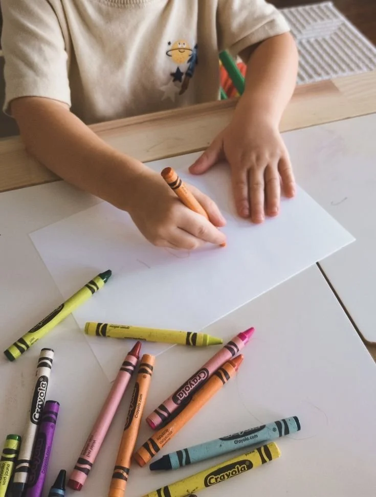 Child drawing on white paper with crayons scattered on table.