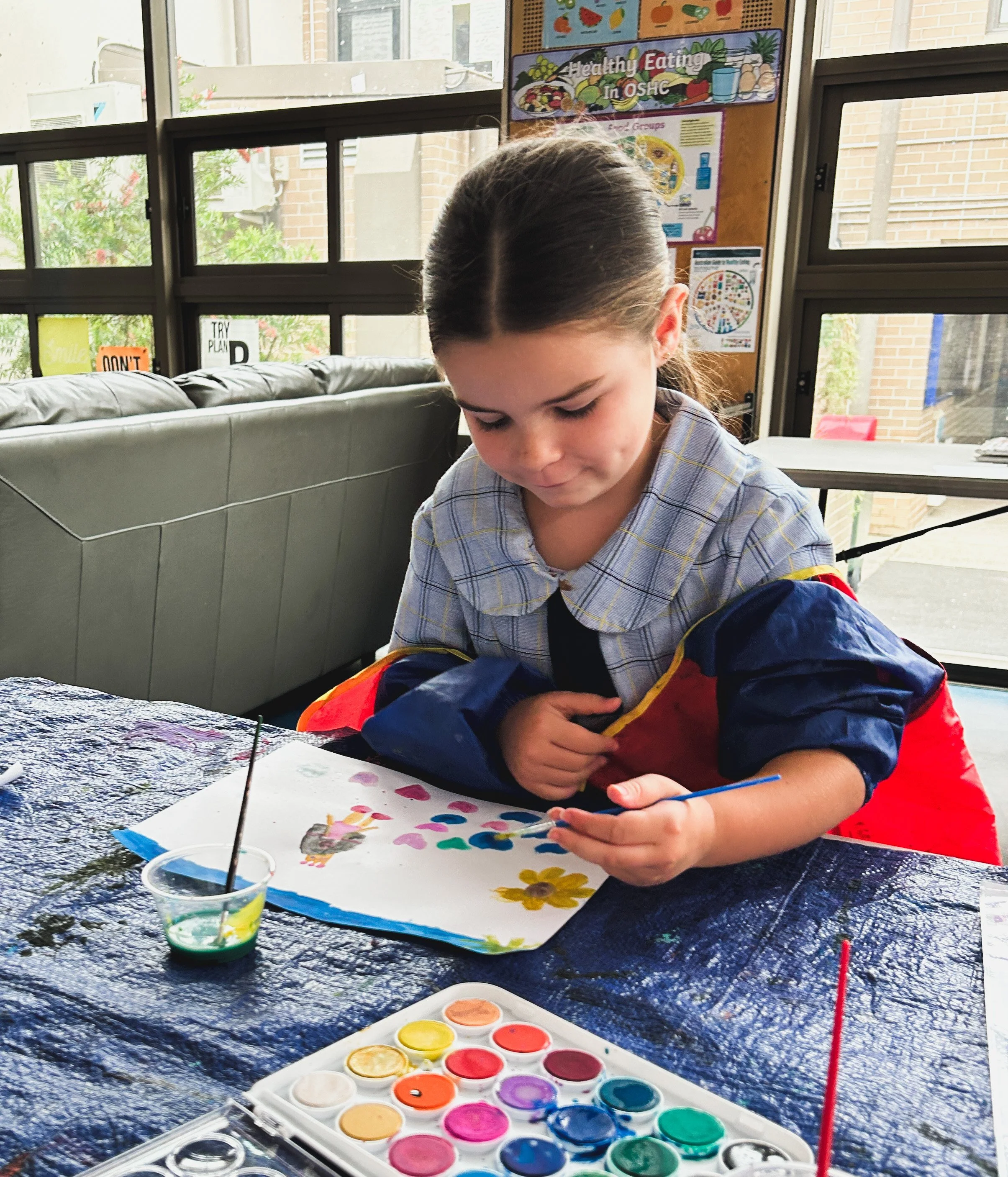 A young girl painting with watercolor paints at a table covered with a dark cloth. She is holding a paintbrush and appears to be focused on her artwork. There are containers of watercolor paints and a paper with painted flowers and hearts on the tabl