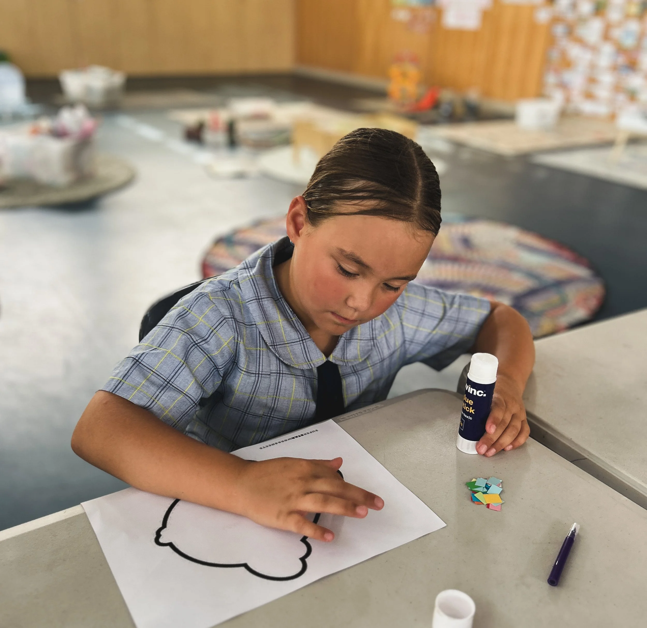 Close-up of a boy with wet hair working on a project at a classroom desk, with colorful paper pieces and a glue stick nearby, and a blurred classroom background.