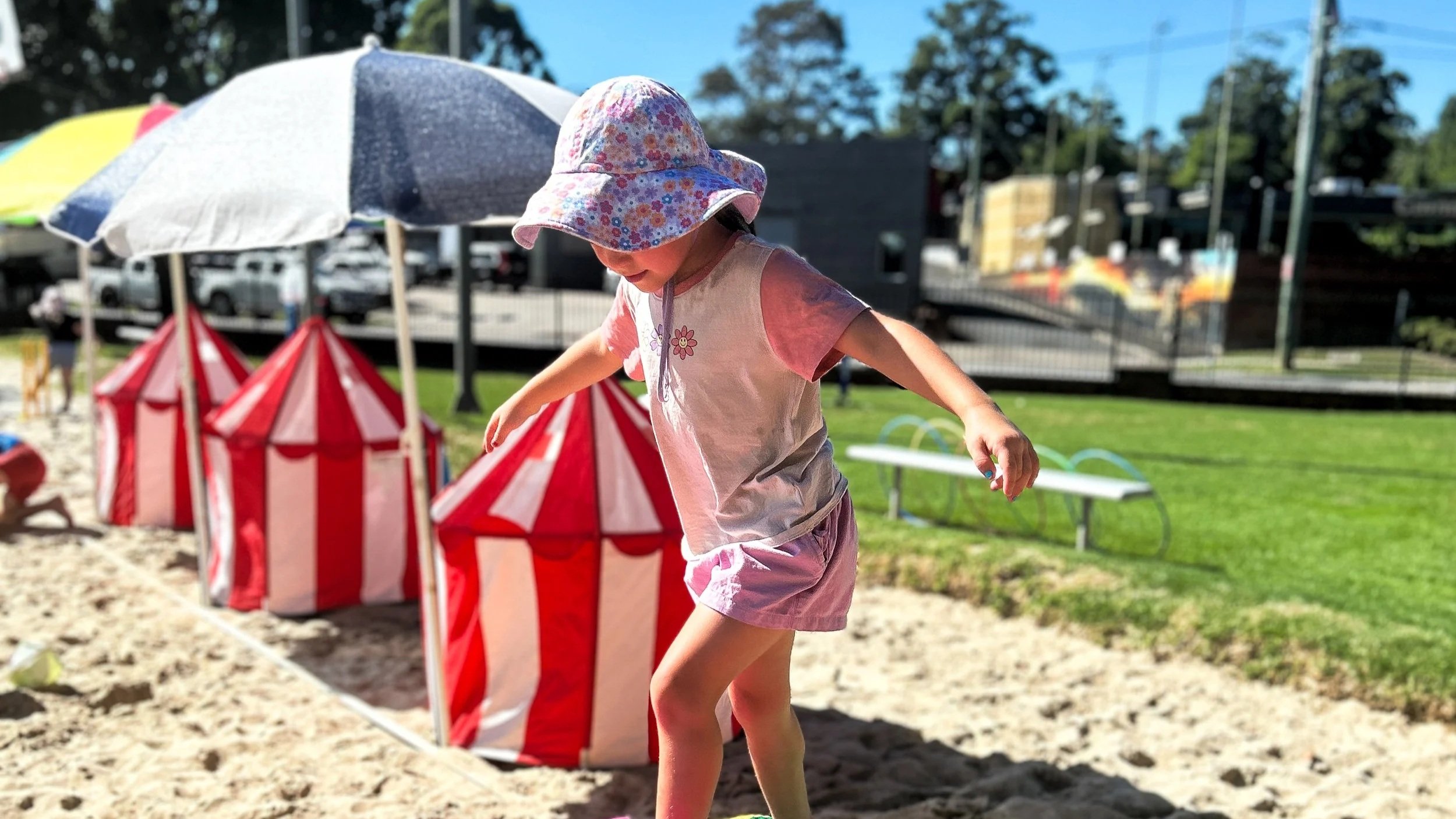 A young girl wearing a floral hat and pink shorts playing at a beach with striped beach tents and umbrellas in the background on a sunny day.