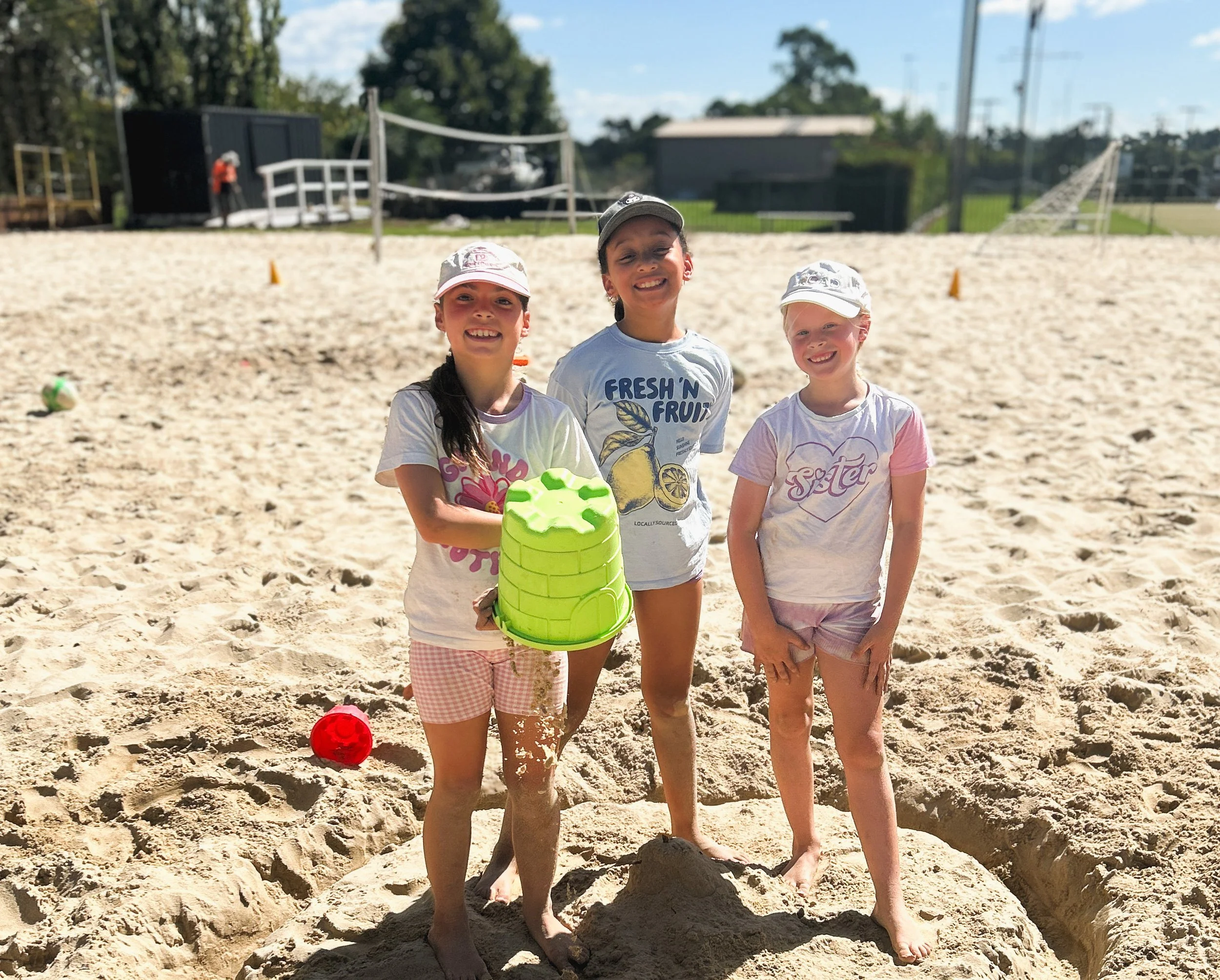 Three young girls smiling on a sandy beach, holding a yellow plastic container, with soccer balls nearby. They are dressed in casual summer clothes and hats, and are standing in front of a sandy area with some playground structures and trees in the b