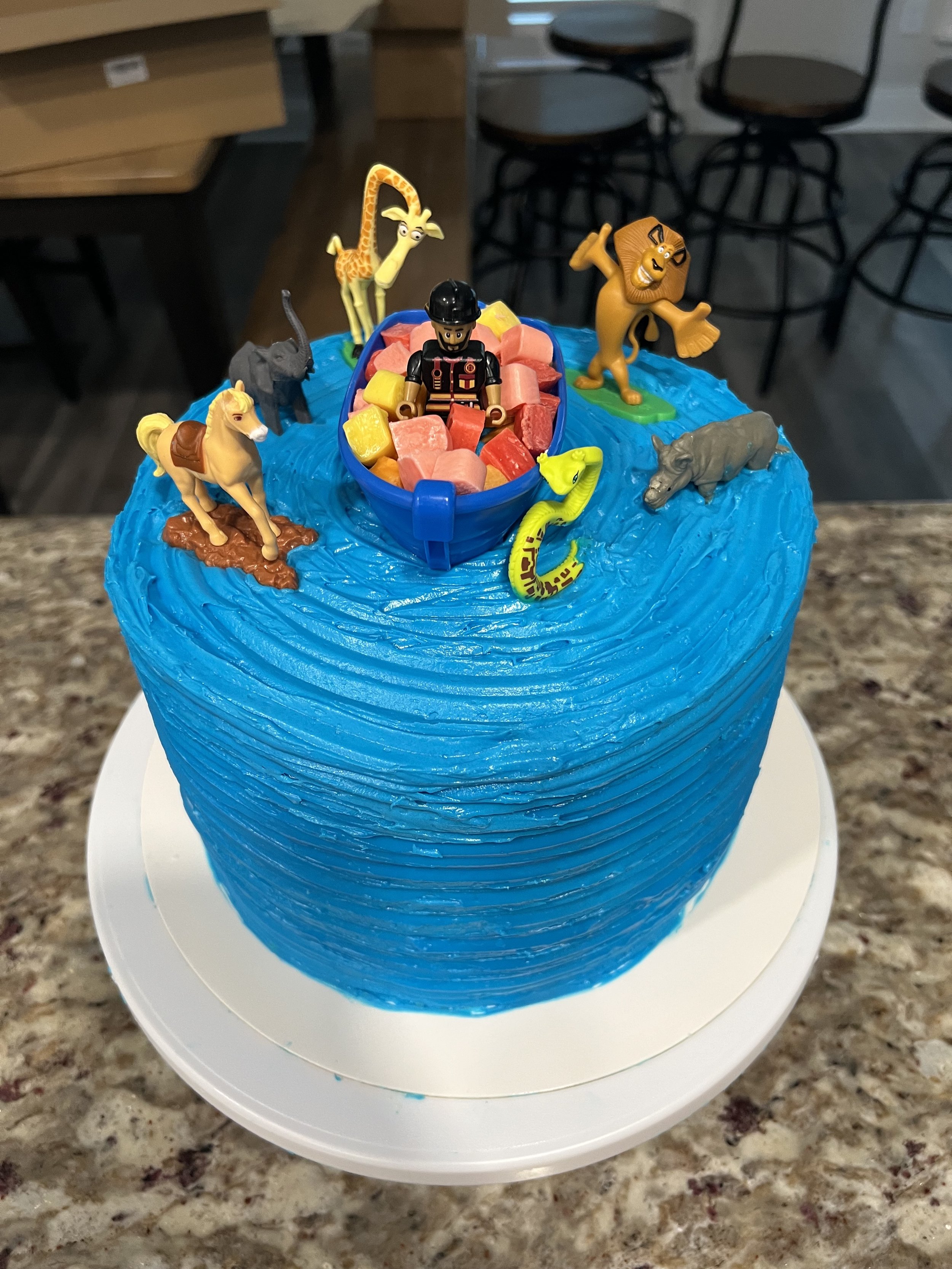 A birthday cake decorated with blue frosting and toy figurines from the movie Madagascar, including Alex the lion, Marty the zebra, Melman the giraffe, Gloria the hippo, and a toy of a zookeeper in a boat with fruit snacks.