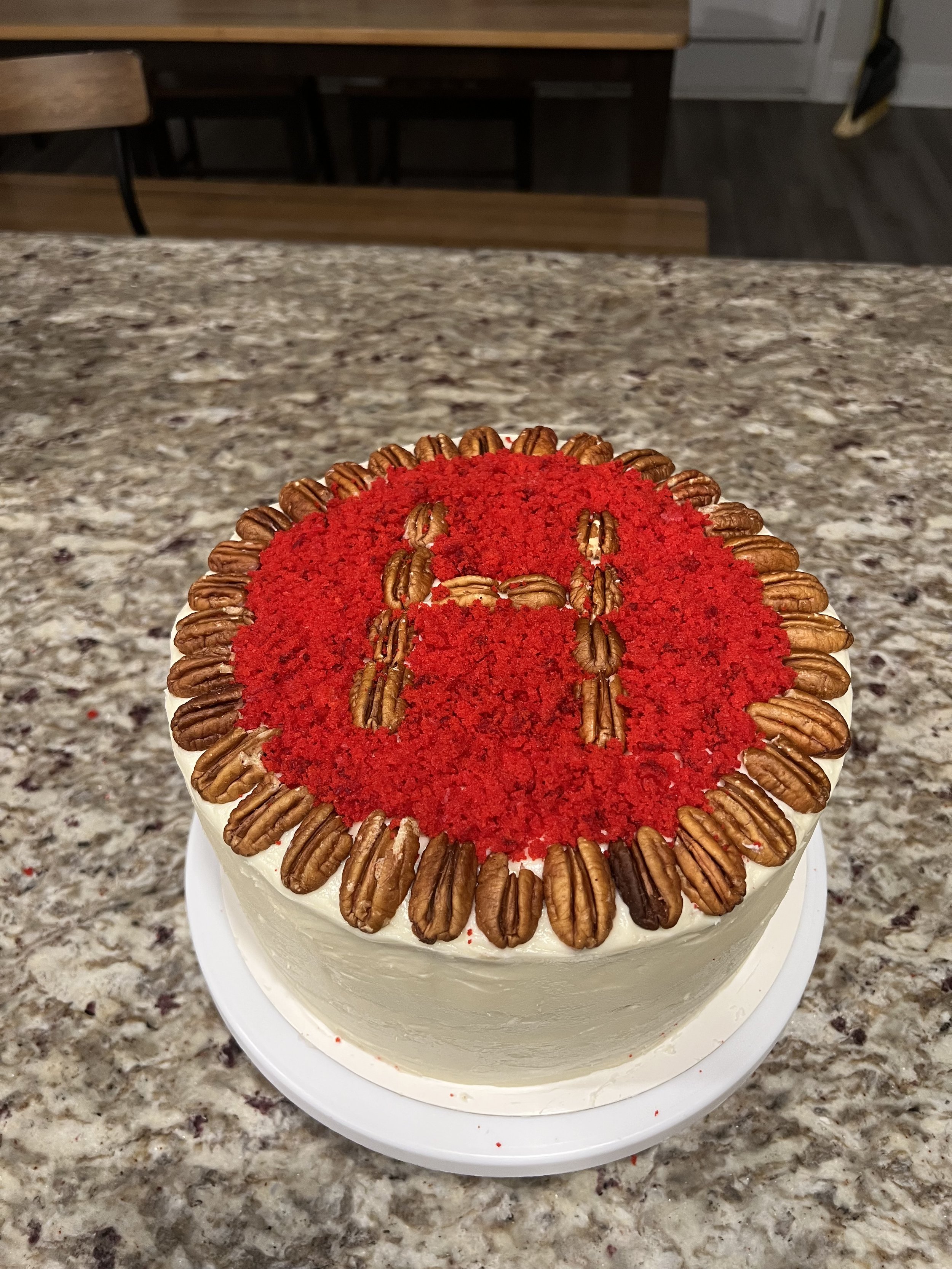 A round cake with white frosting, decorated with pecans around the edges and red cake crumbs on top. 
