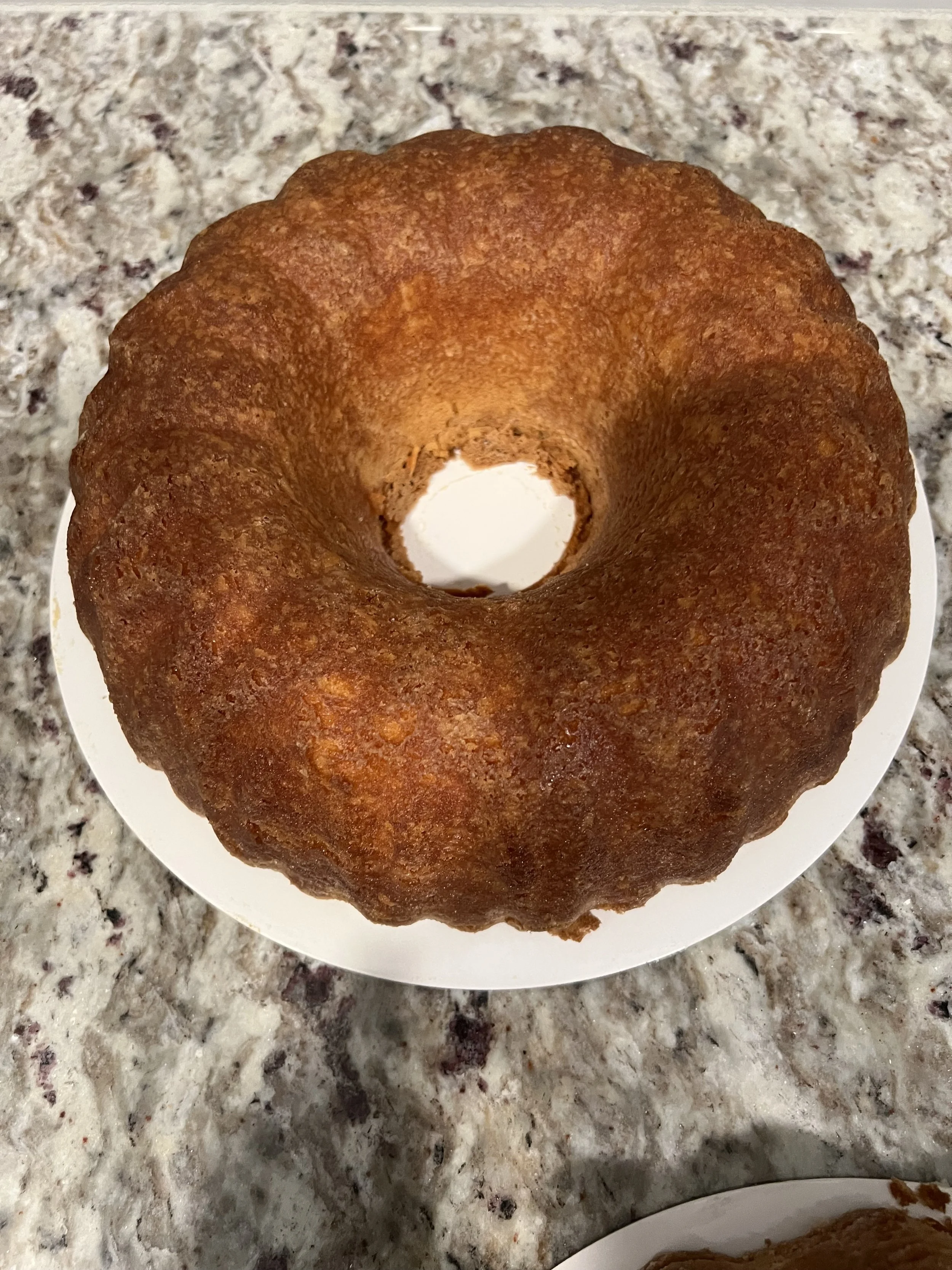 A golden-brown Bundt cake on a white plate sitting on a speckled granite countertop.