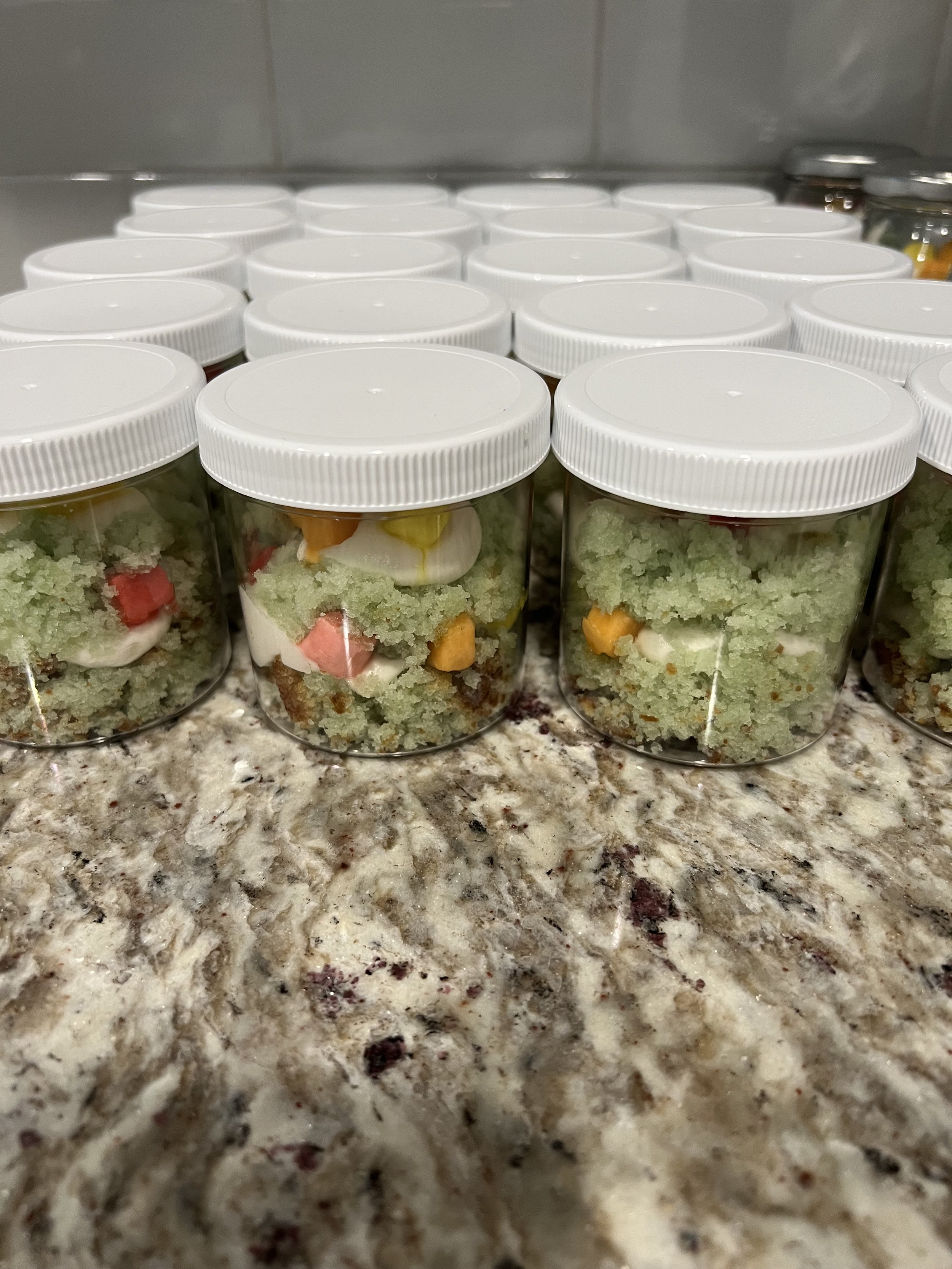 Multiple small jars with white lids, filled with a layered dessert containing green, red, white, and orange ingredients, placed on a speckled granite countertop.