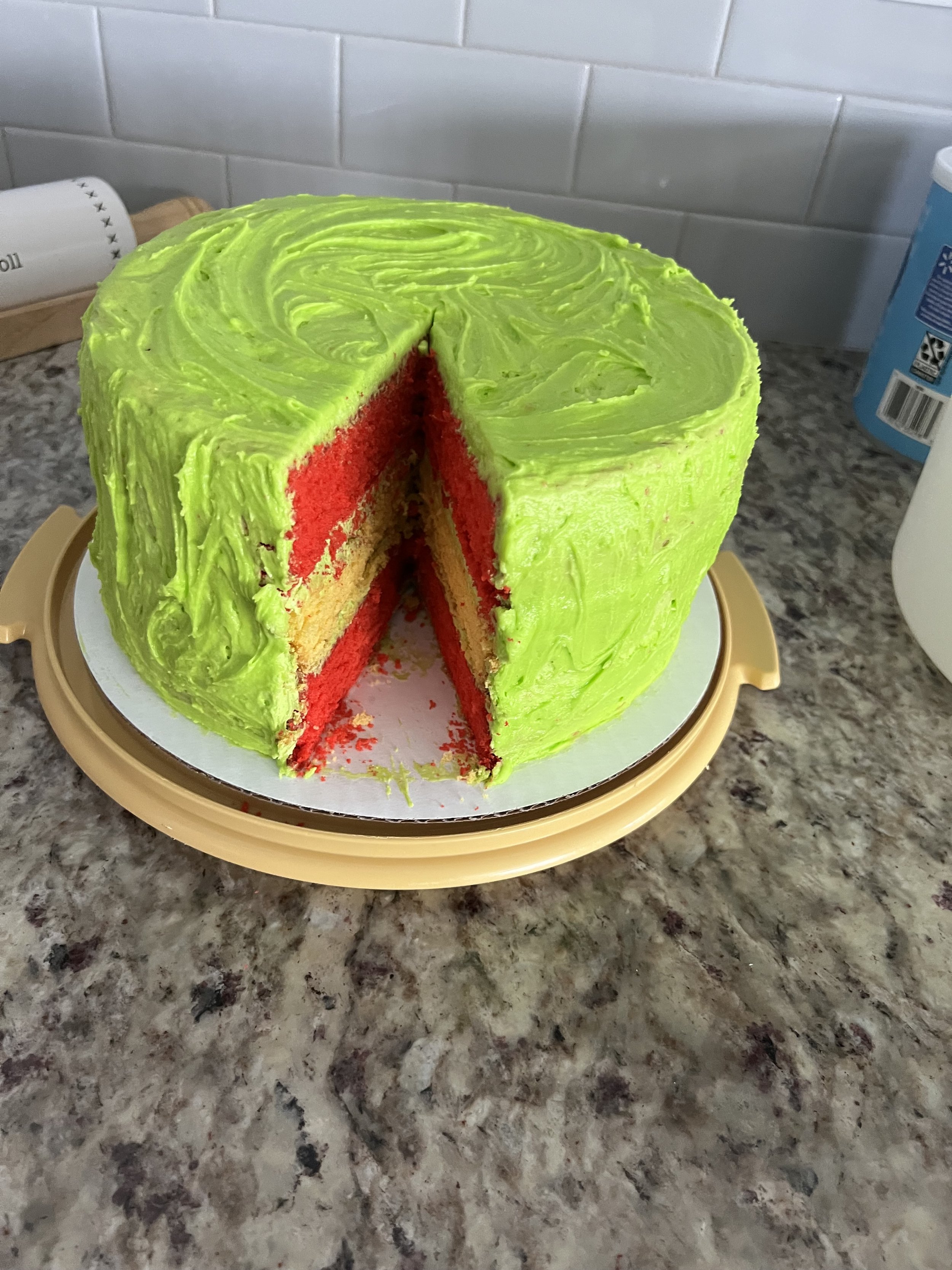A colorful layered cake with bright green frosting on the outside and red and yellow layers inside, placed on a cake stand on a kitchen countertop.