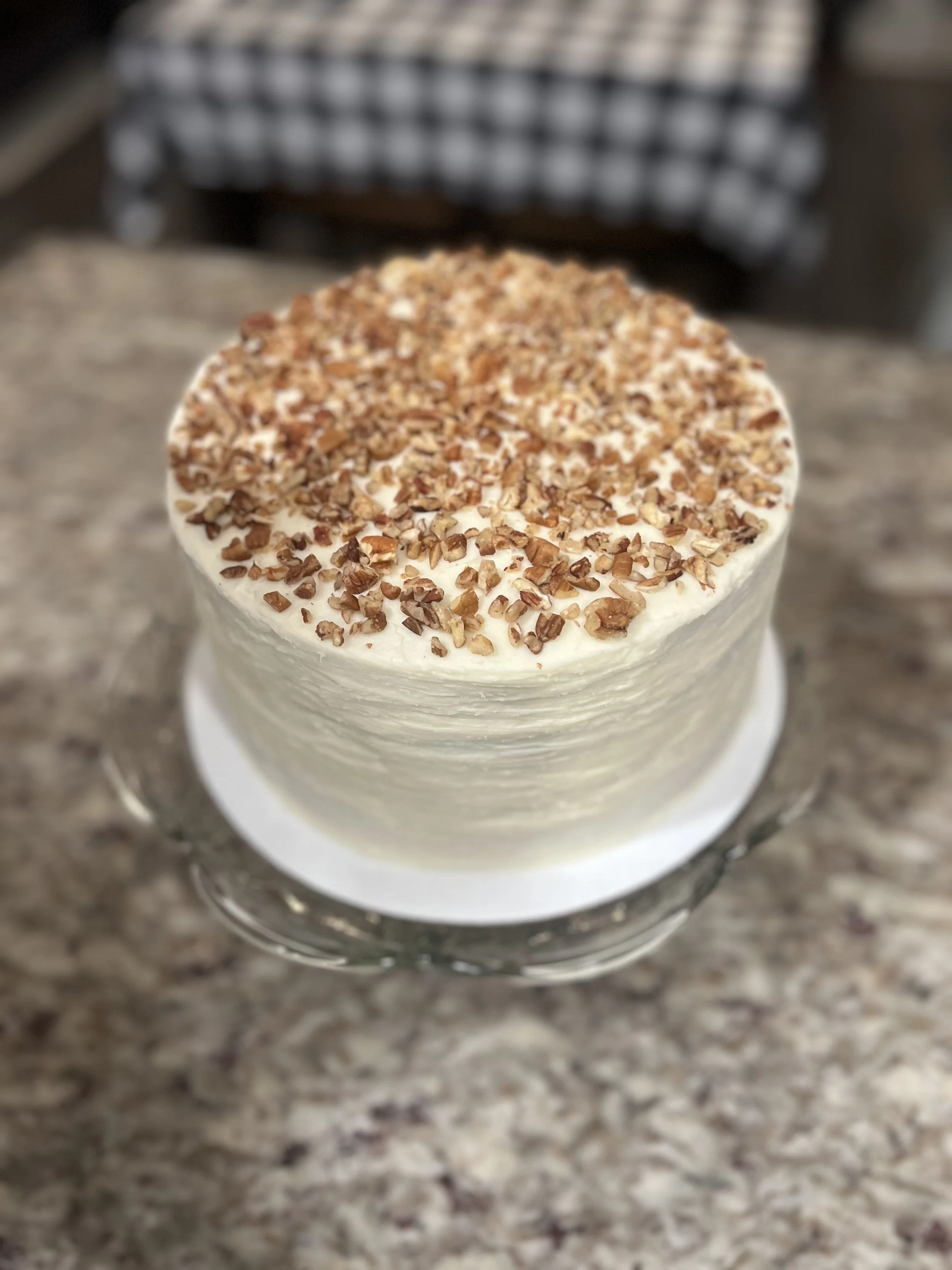 A cake with white frosting topped with chopped nuts on a glass cake stand, placed on a marble countertop.