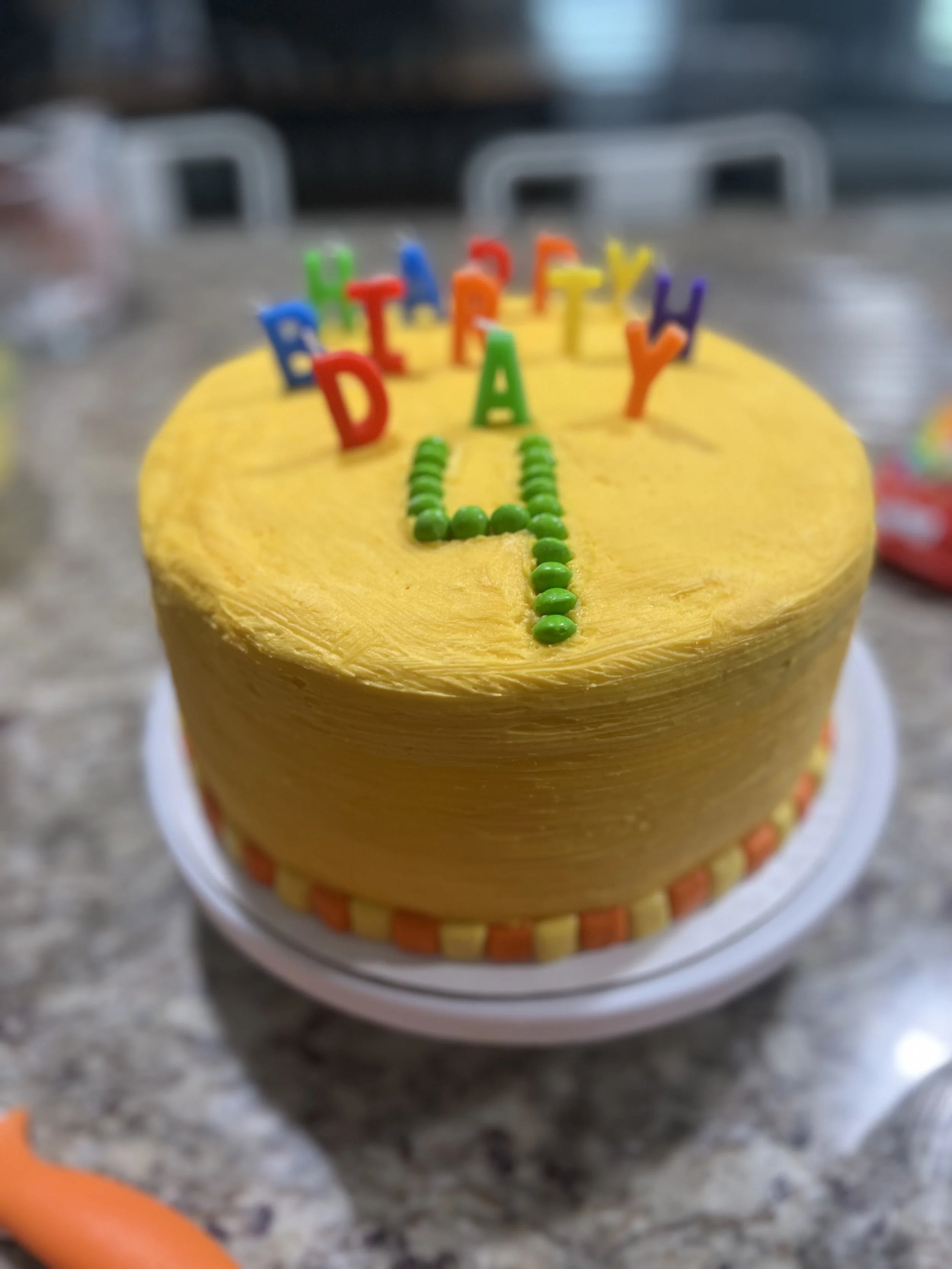 Yellow birthday cake with colorful candle letters spelling 'HAPPY BIRTHDAY' on top, green frosting dots in a line, and orange and yellow decorative border at the bottom, on a gray countertop.