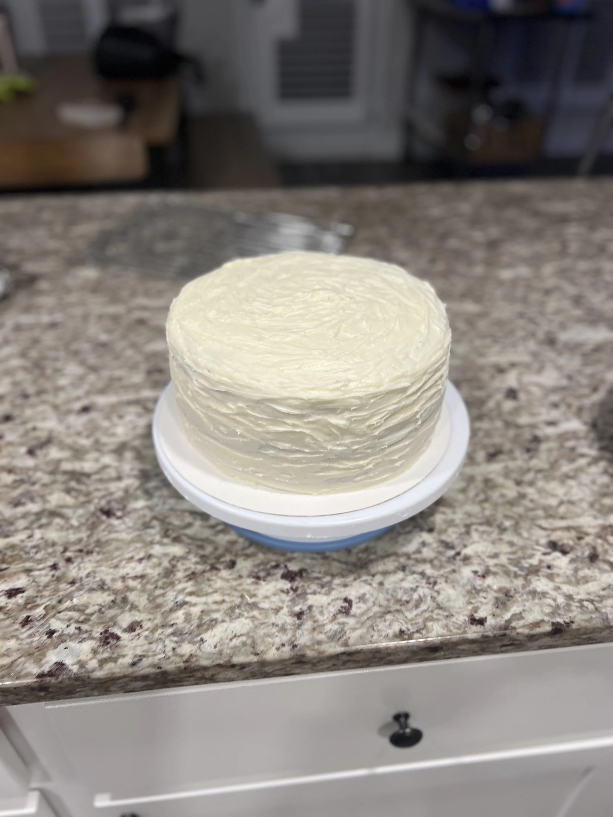A round cake with white frosting on a cake stand, placed on a speckled granite countertop in a kitchen.