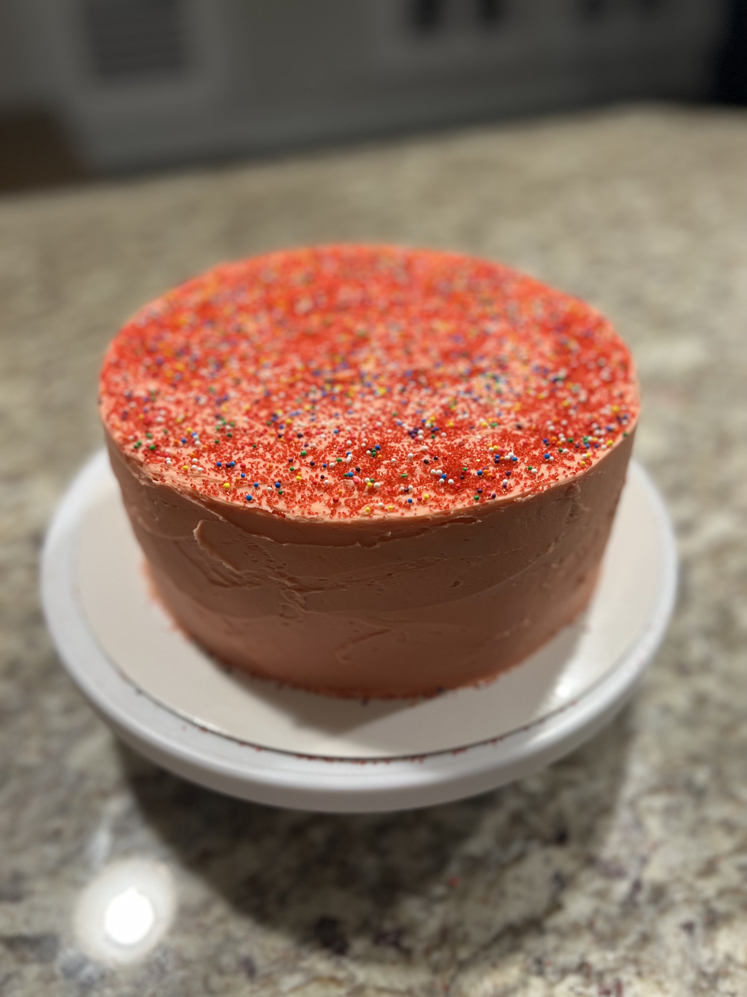 A small round cake with red icing and colorful sprinkles on top, placed on a white plate on a granite countertop.