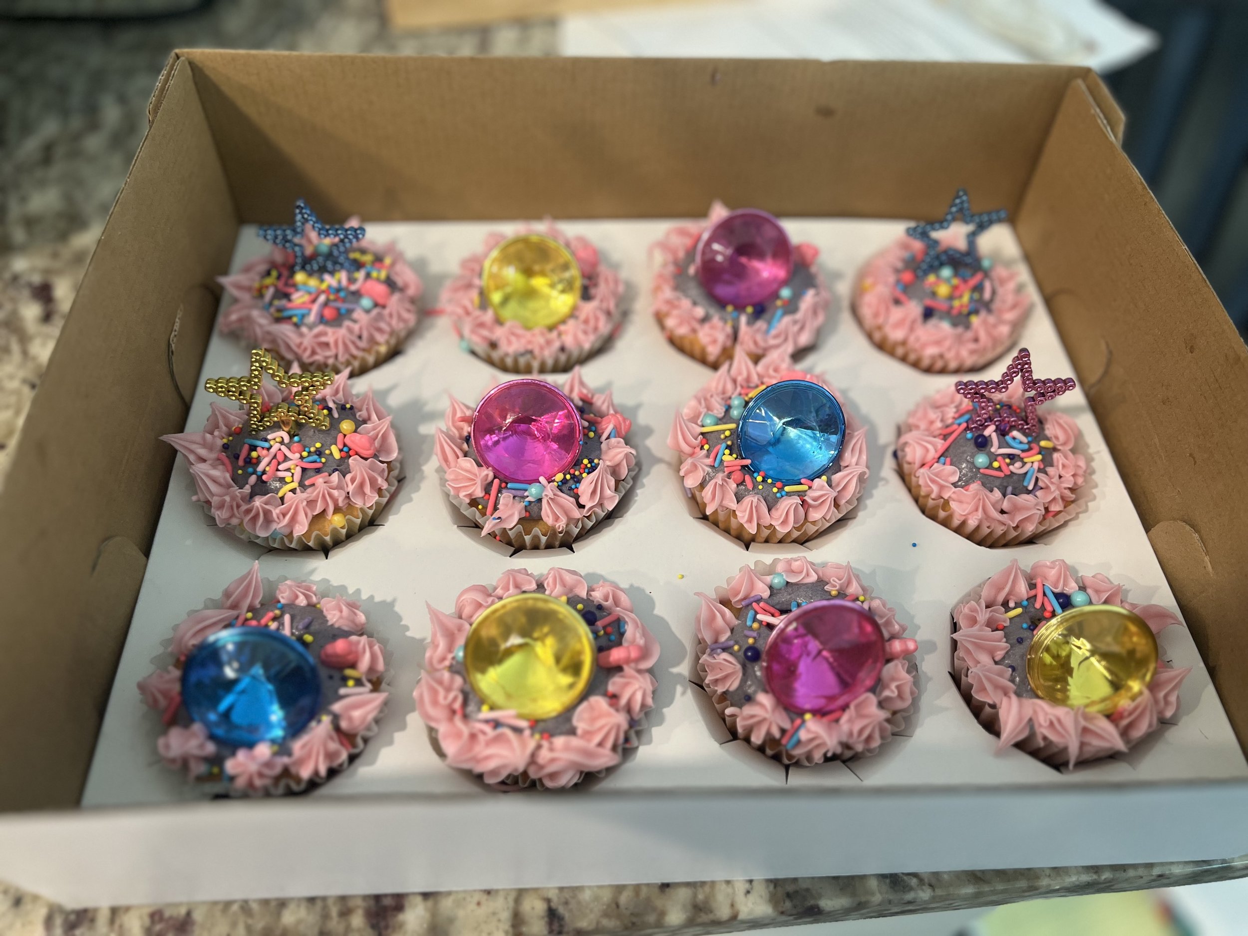 Box of decorated cupcakes with pink frosting, colorful sprinkles, star-shaped decorations, and shiny gem-like toppers in yellow, pink, and blue colors.