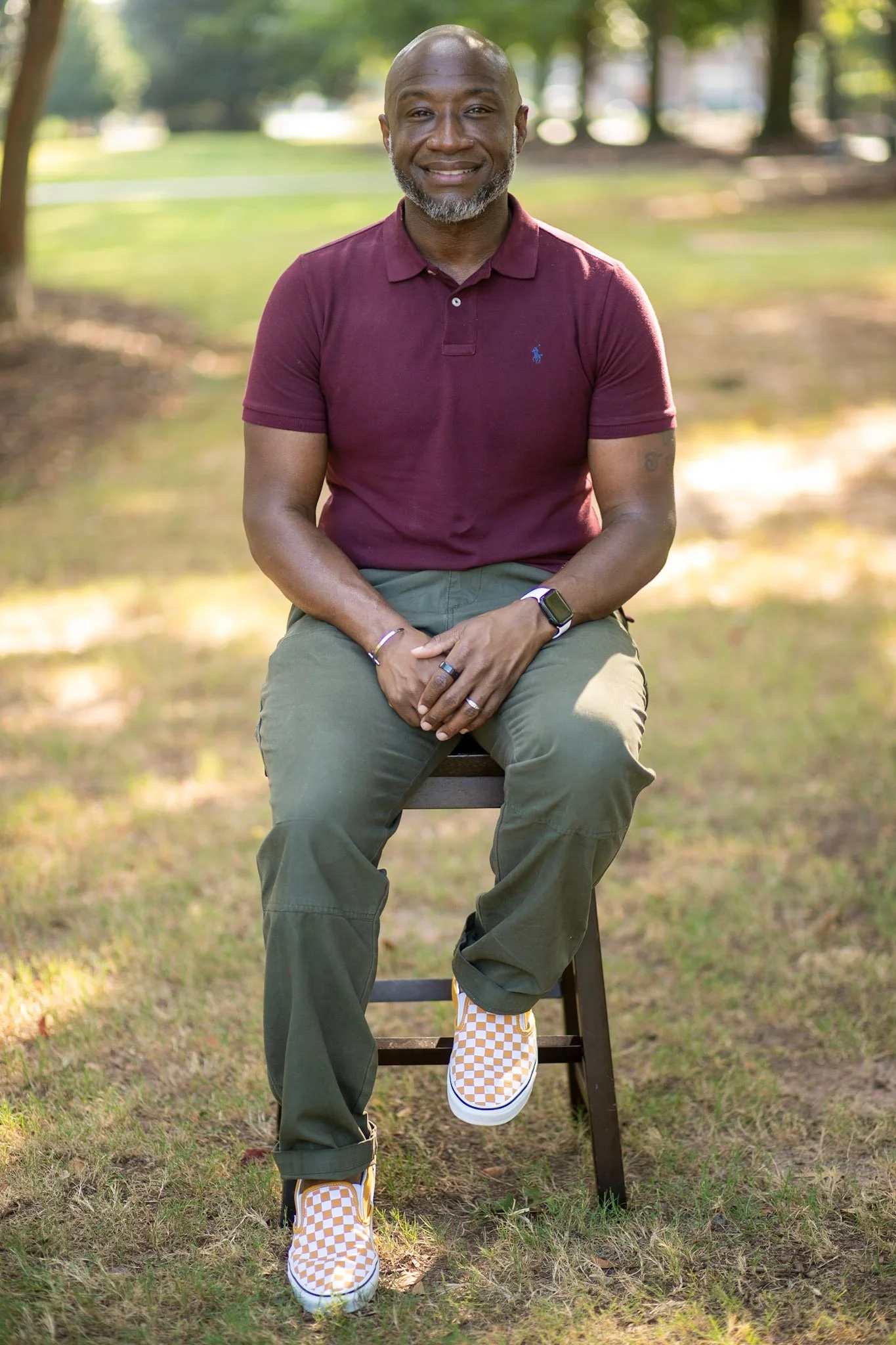 A middle-aged African American man with a beard, sitting on a wooden stool outdoors in a park, smiling at the camera, wearing a maroon polo shirt, green cargo pants, and checkered yellow and white sneakers, with trees in the background and sunlight filtering through the leaves.