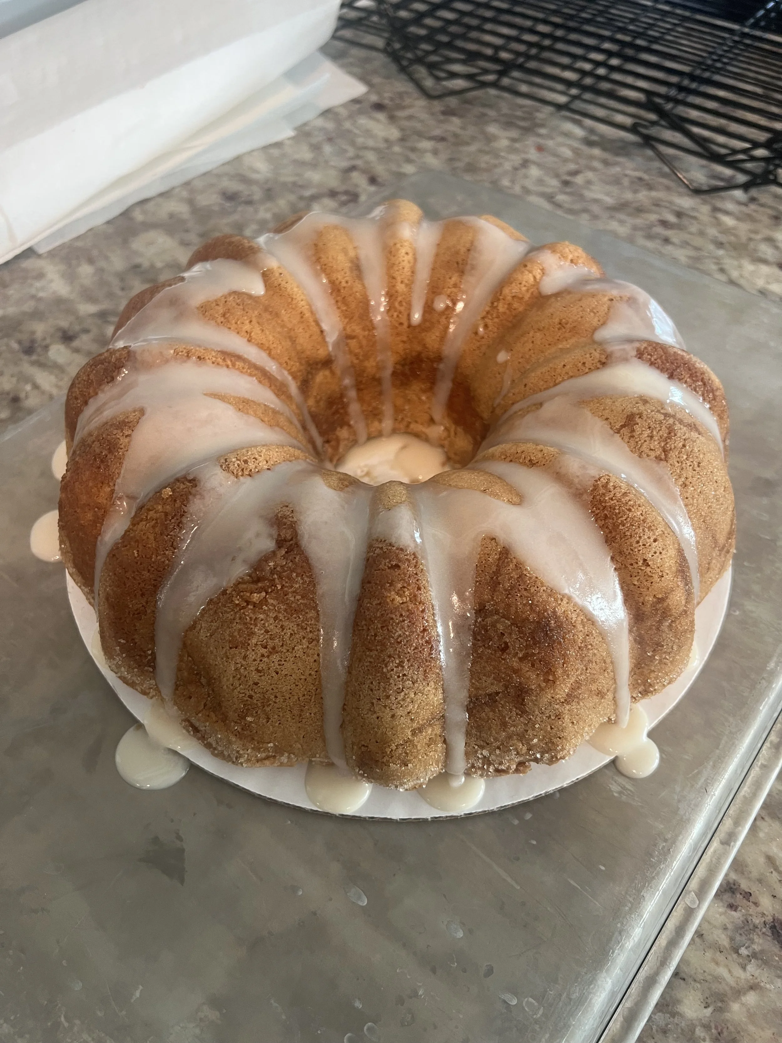 Bundt cake with white glaze drip on top, resting on a metal surface with a cooling rack in the background.