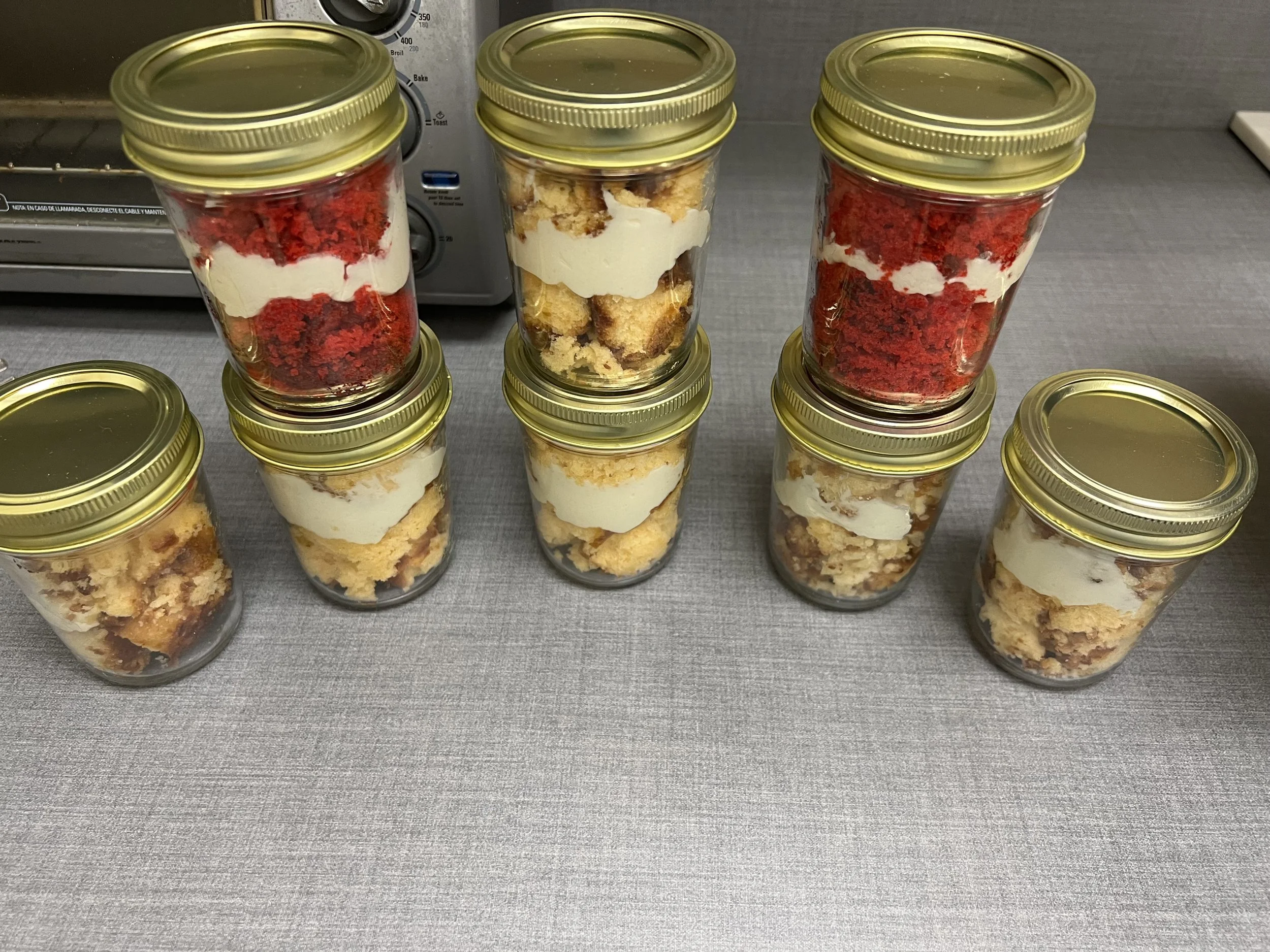 Six glass jars with gold lids filled with layers of red velvet cake, vanilla cream, and cake crumbs, arranged on a gray counter with a toaster oven in the background.