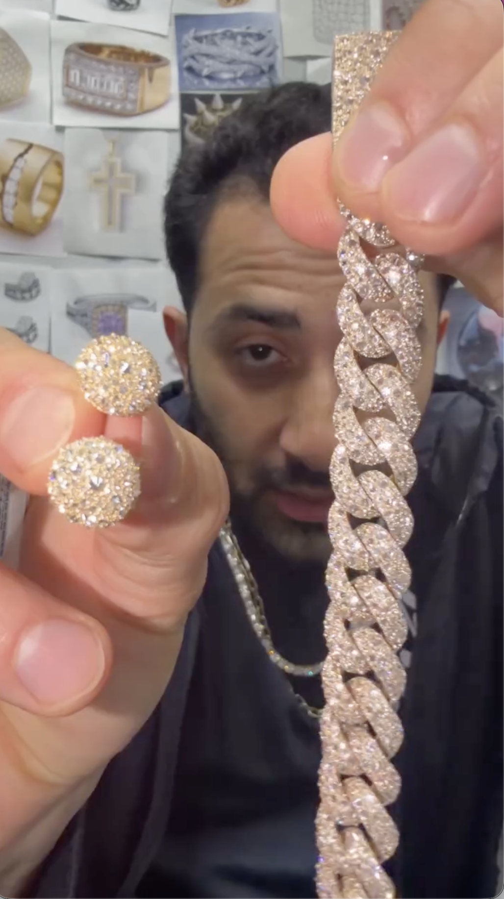 TraxNYC holds up a diamond ring, earrings, and a Cuban link, surrounded by jewelry display images on the wall.