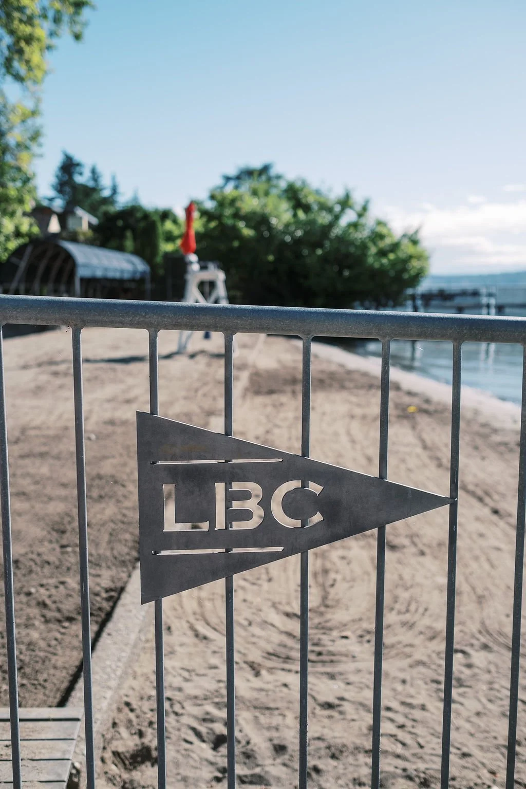A metal gate with the letters 'LBC' cut out, blocking a sandy beach area with trees, a structure, and a red object in the background near water.