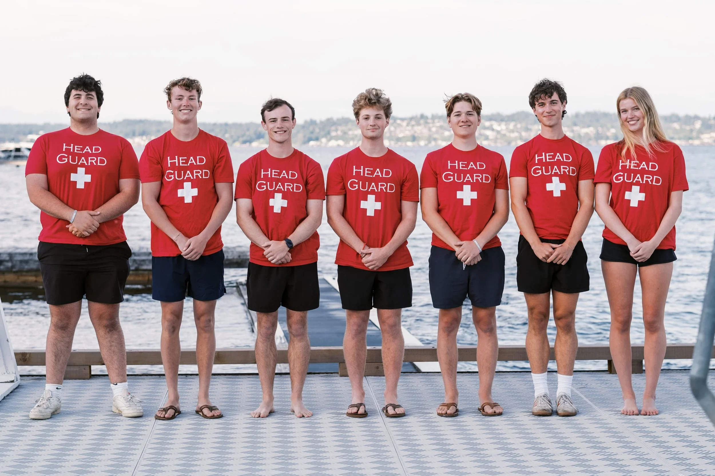 A group of seven young people standing on a dock near water, wearing red t-shirts with 'Head Guard' and a white cross, smiling and posing for the photo with a shoreline with buildings in the background.