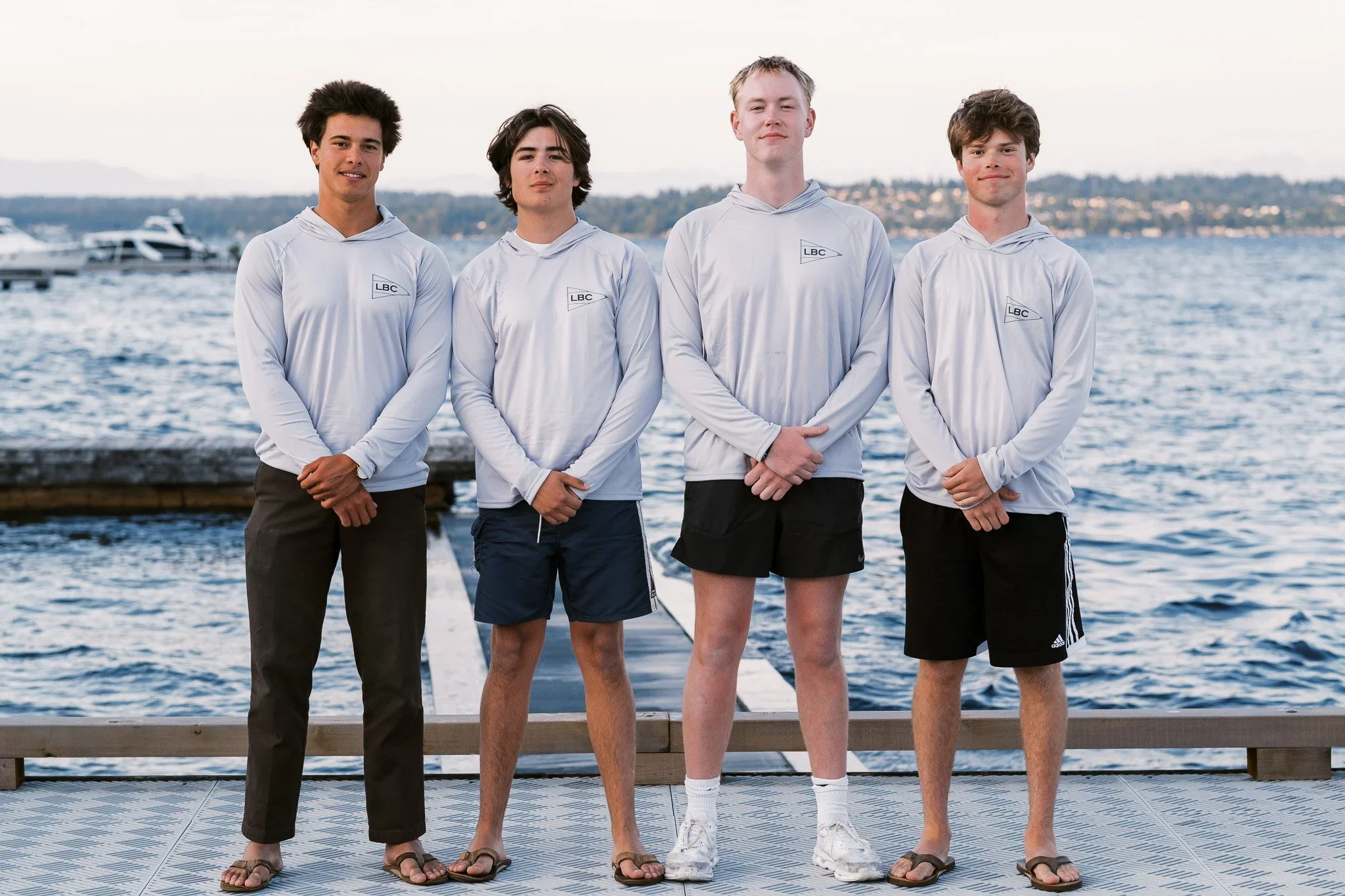 Four teenage boys standing on a dock near the water with boats in the background, wearing white hooded shirts with a logo, shorts, and flip-flops.
