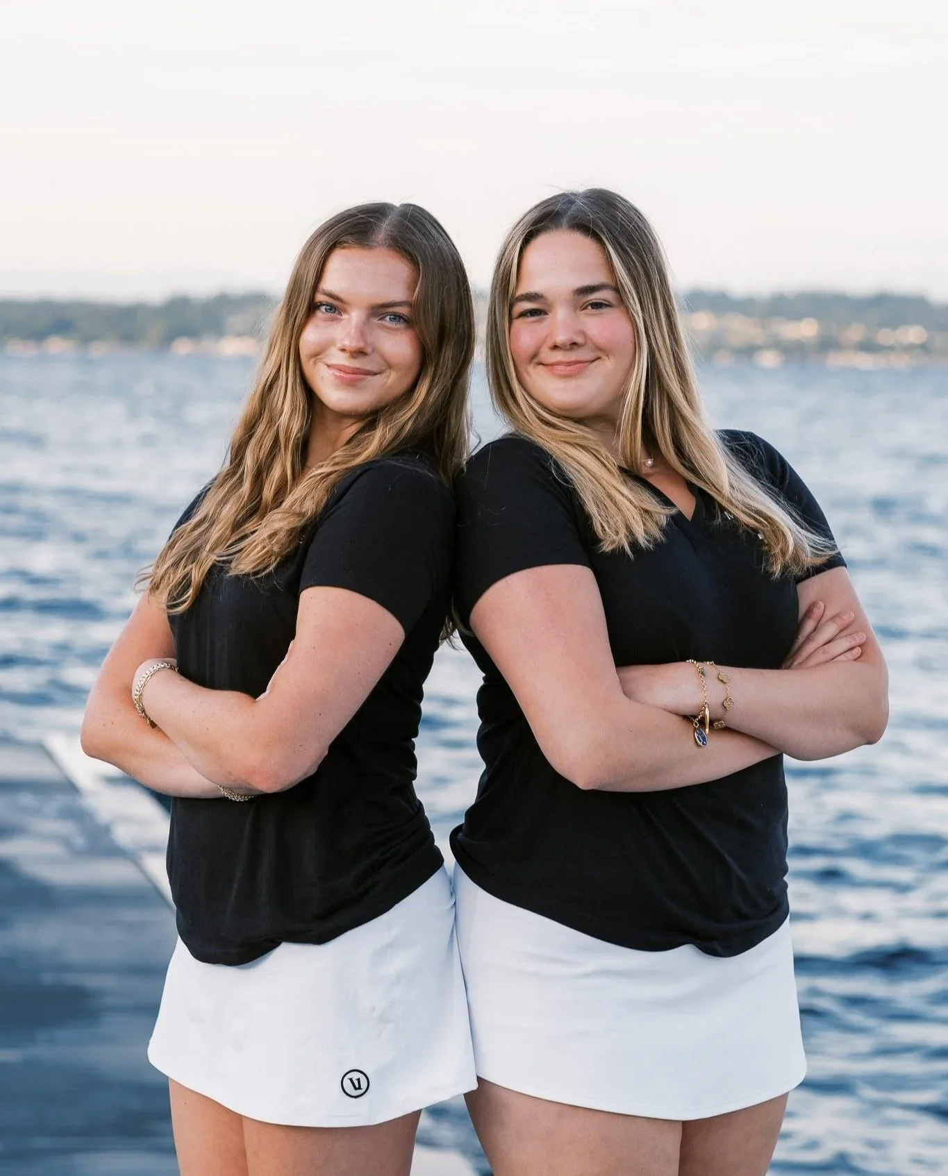 Two women standing back to back with arms crossed in front of a body of water, smiling, wearing black shirts and white shorts, with a distant shoreline in the background.