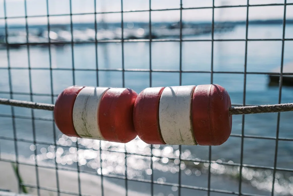 A close-up of a buoy attached to a wire fence near a body of water with boats in the background.