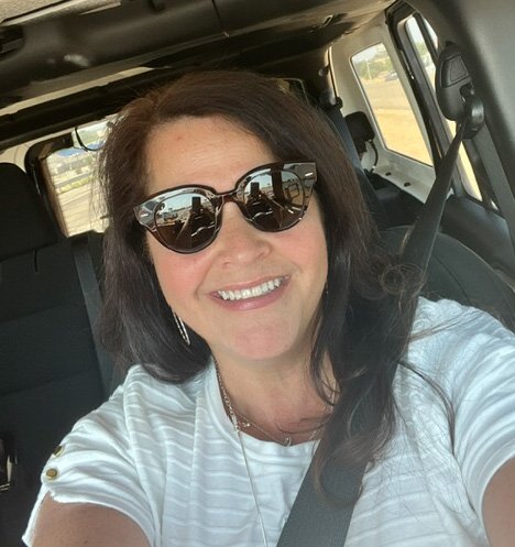 Woman with dark hair wearing sunglasses and a white shirt, smiling, sitting in a car.
