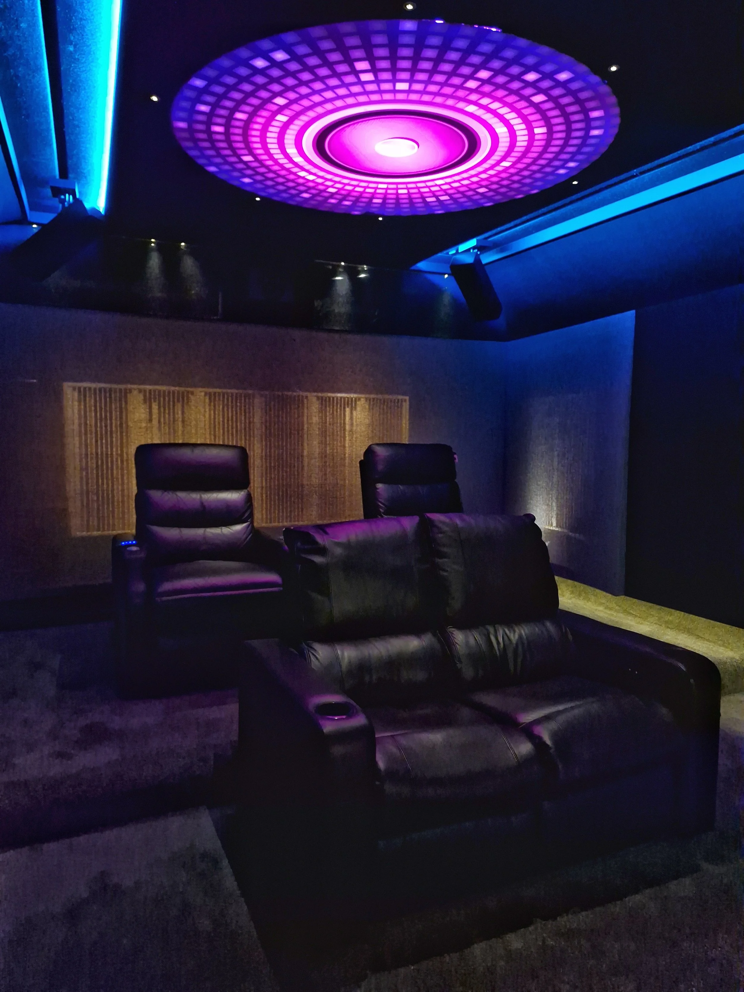 Dark theater room with black leather reclining chairs, purple lighting, and a large circular purple and pink ceiling light feature.