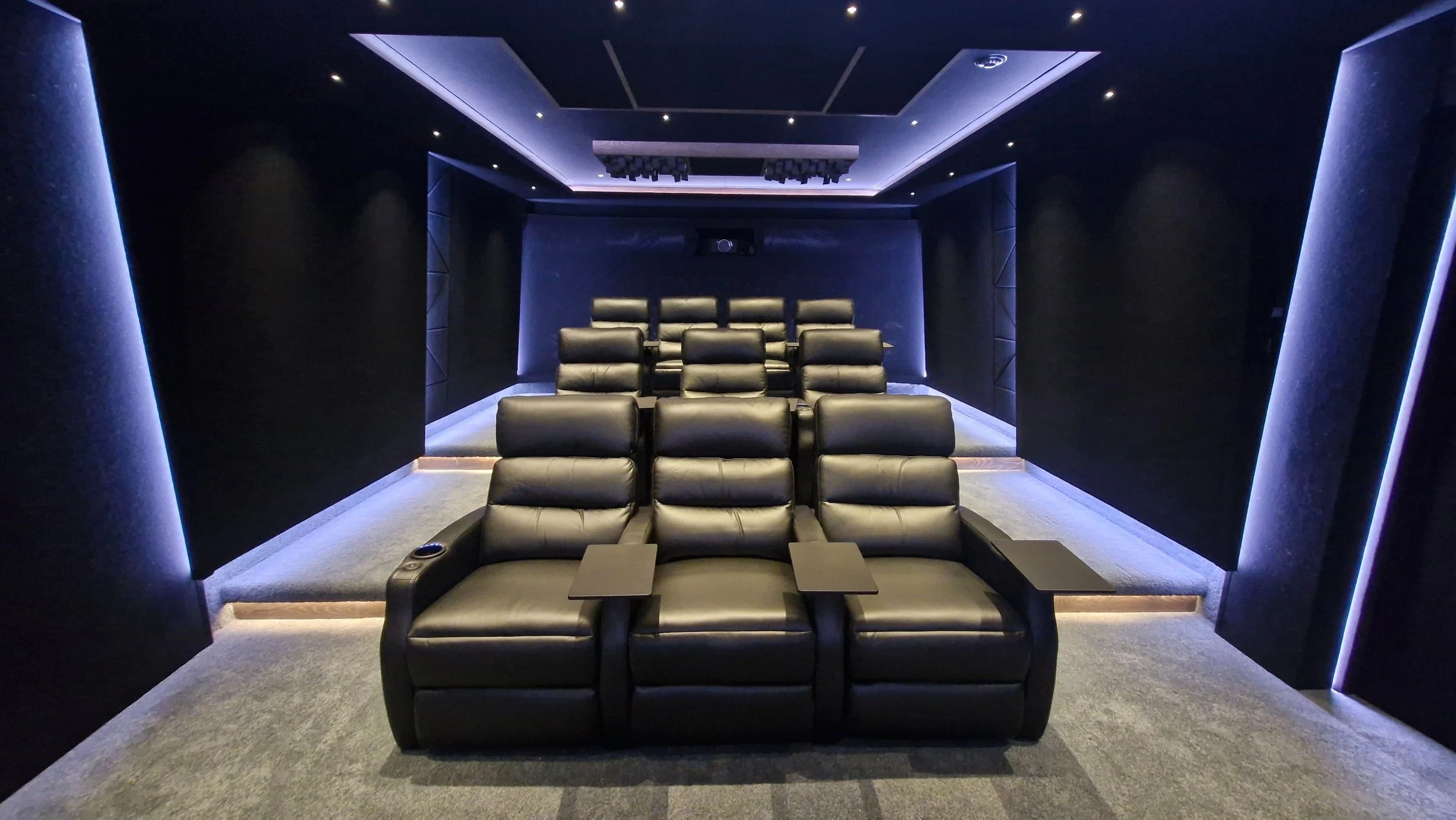 A modern home theater room with black leather recliner seats arranged in rows and a large screen on the back wall. The room features dark walls and ceiling with ambient blue lighting.