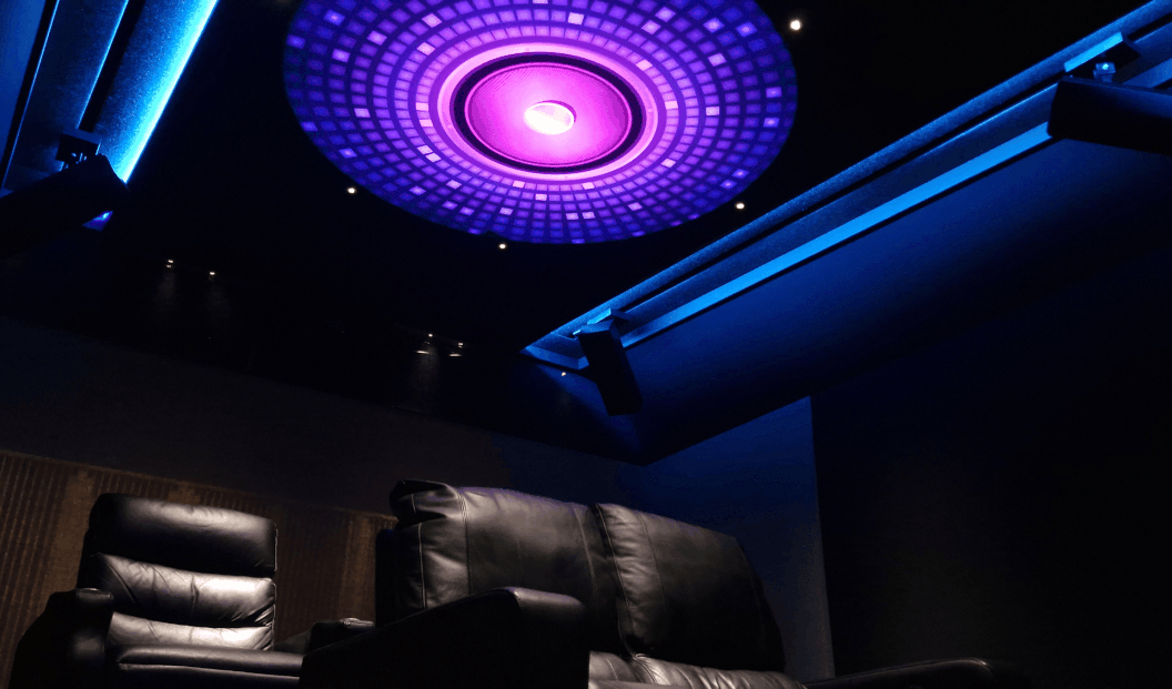 Dimly lit home theater with black leather recliners and a ceiling featuring a large circular purple and blue LED light installation.