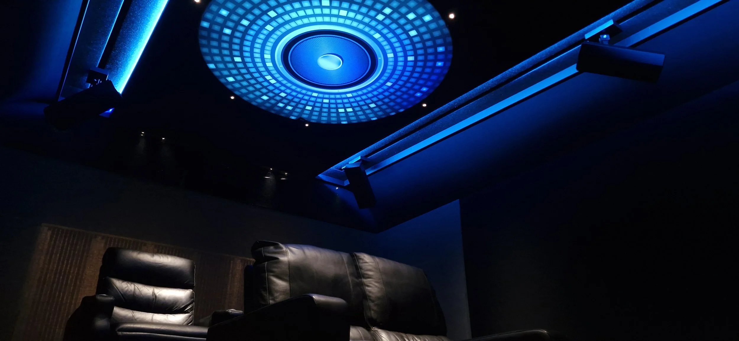 A home theater room with a blue-lit ceiling installation, black leather recliner chairs, and acoustic panels on the walls.
