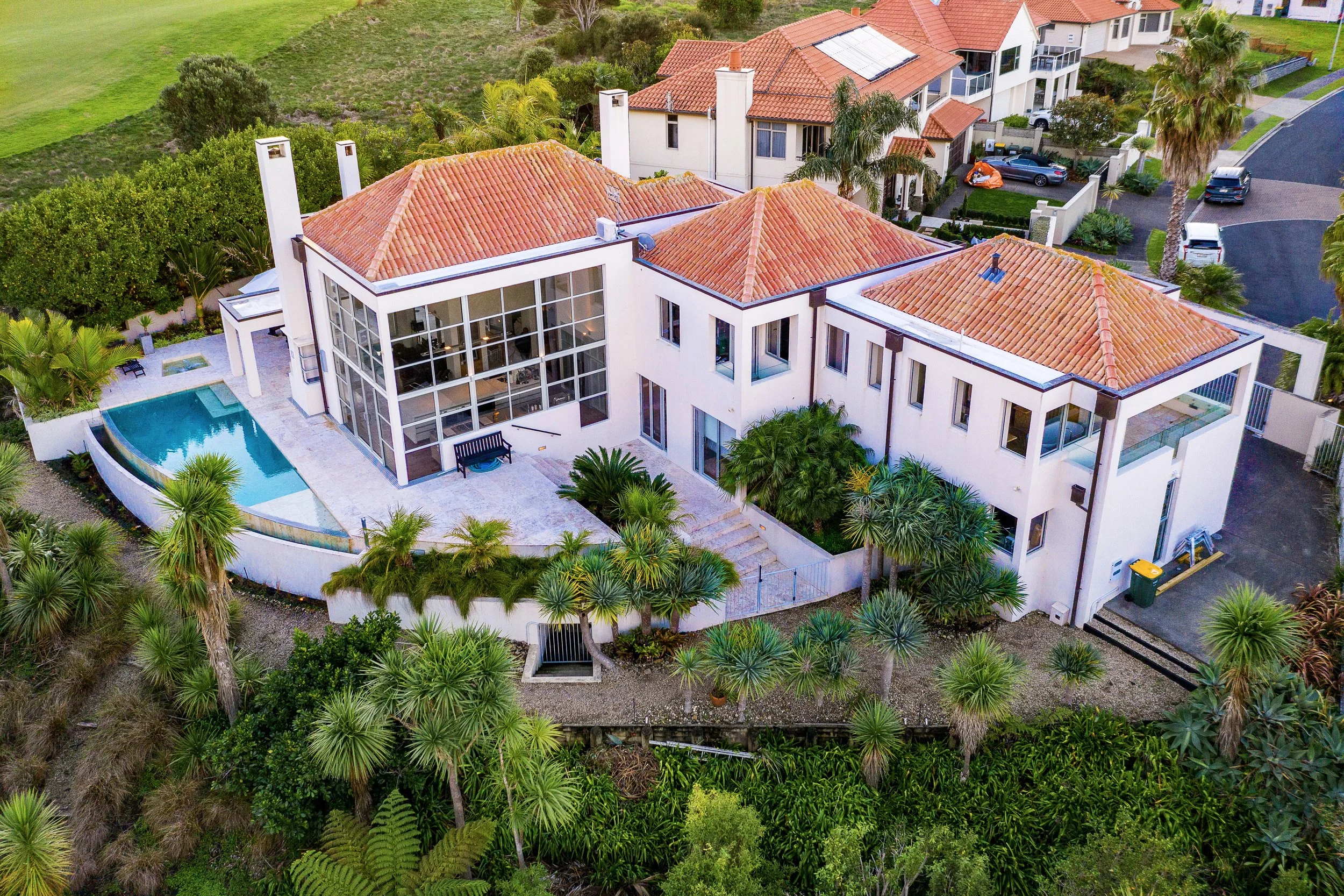 Aerial view of a modern house with white exterior walls and red-tiled roof, surrounded by lush greenery, a swimming pool, and neighboring houses on a residential street.