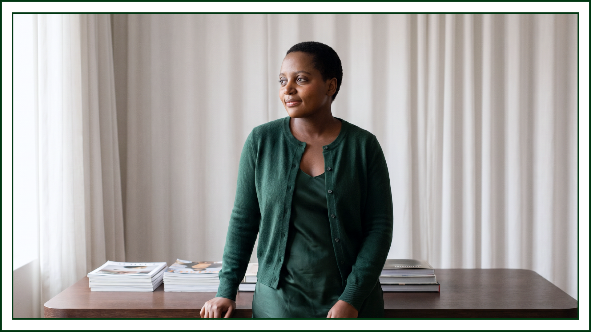 Lillian Ingabire, editorial designer and founder of Studio Kalili, standing beside a wooden table stacked with design books and magazines, looking off to the side.