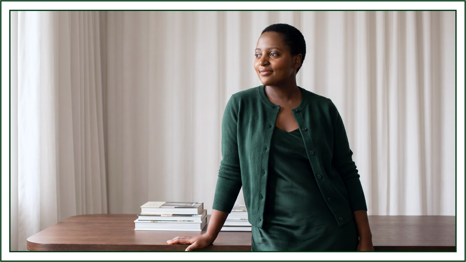 Portrait of Lillian Ingabire of Studio Kalili, a brand identity studio in Chicago, leaning against a wooden desk in a green cardigan and dress with design books stacked beside her.