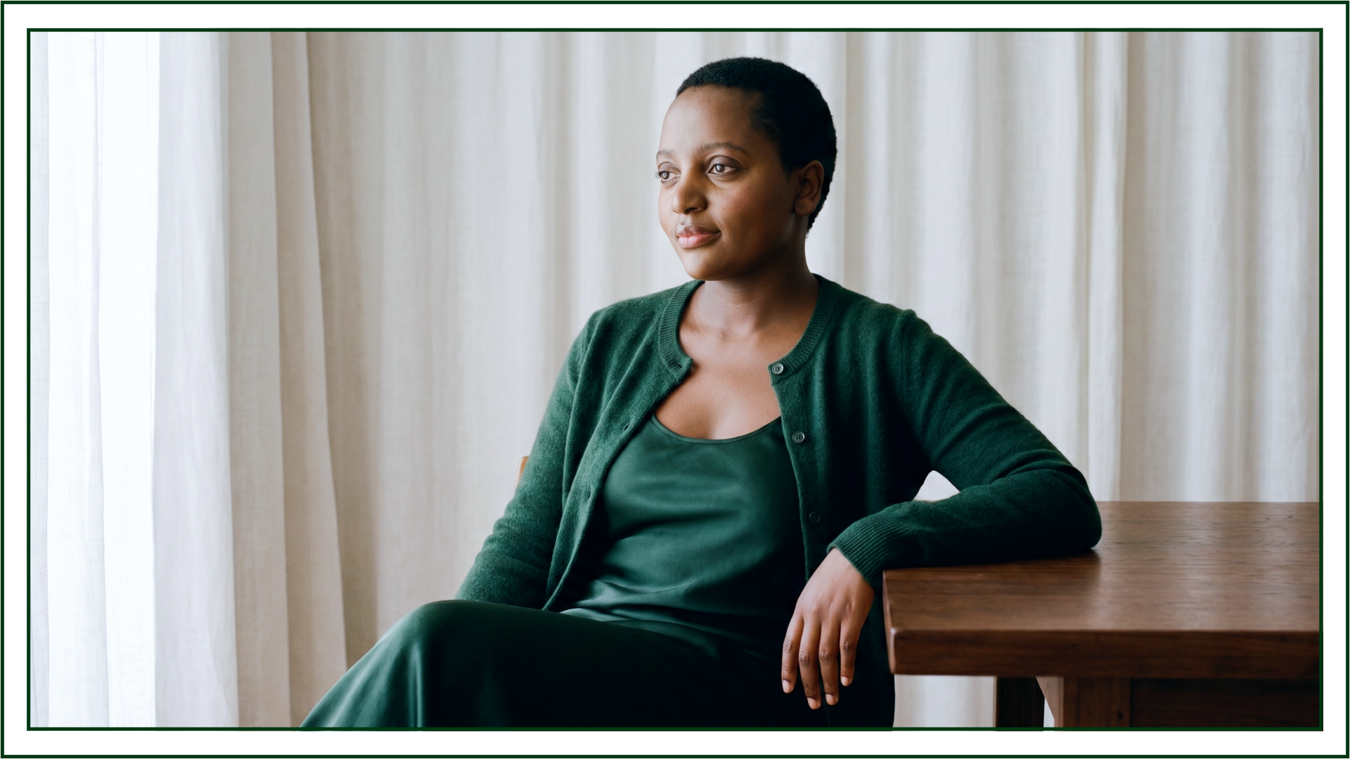 Wide portrait of Lillian Ingabire, editorial brand designer and founder of Studio Kalili, seated at a wooden table in a green cardigan and dress with an arm resting on the table's edge.
