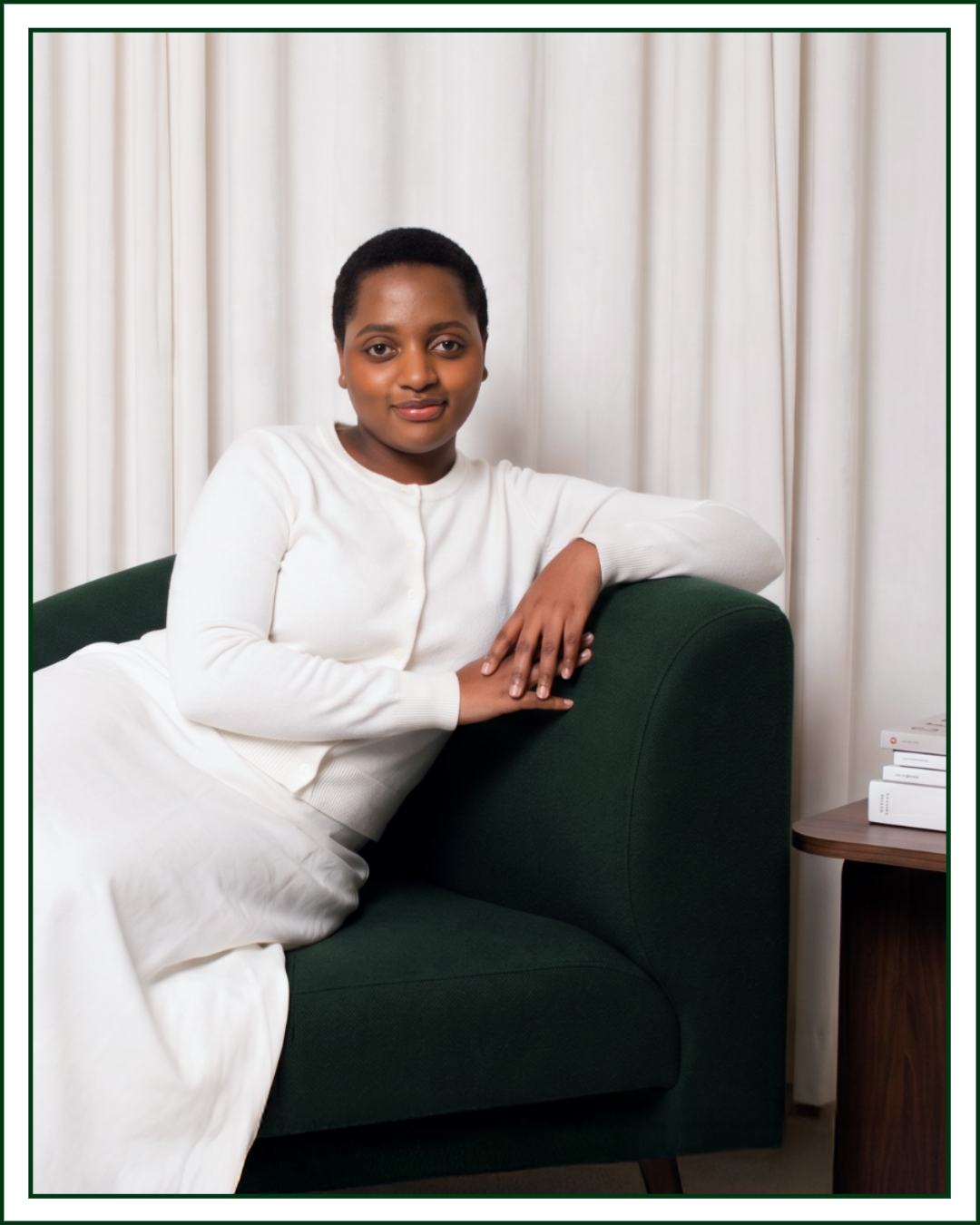 Lillian Ingabire, editorial brand designer and founder of Studio Kalili in Chicago, seated in a green armchair with a stack of design books on a side table beside her.
