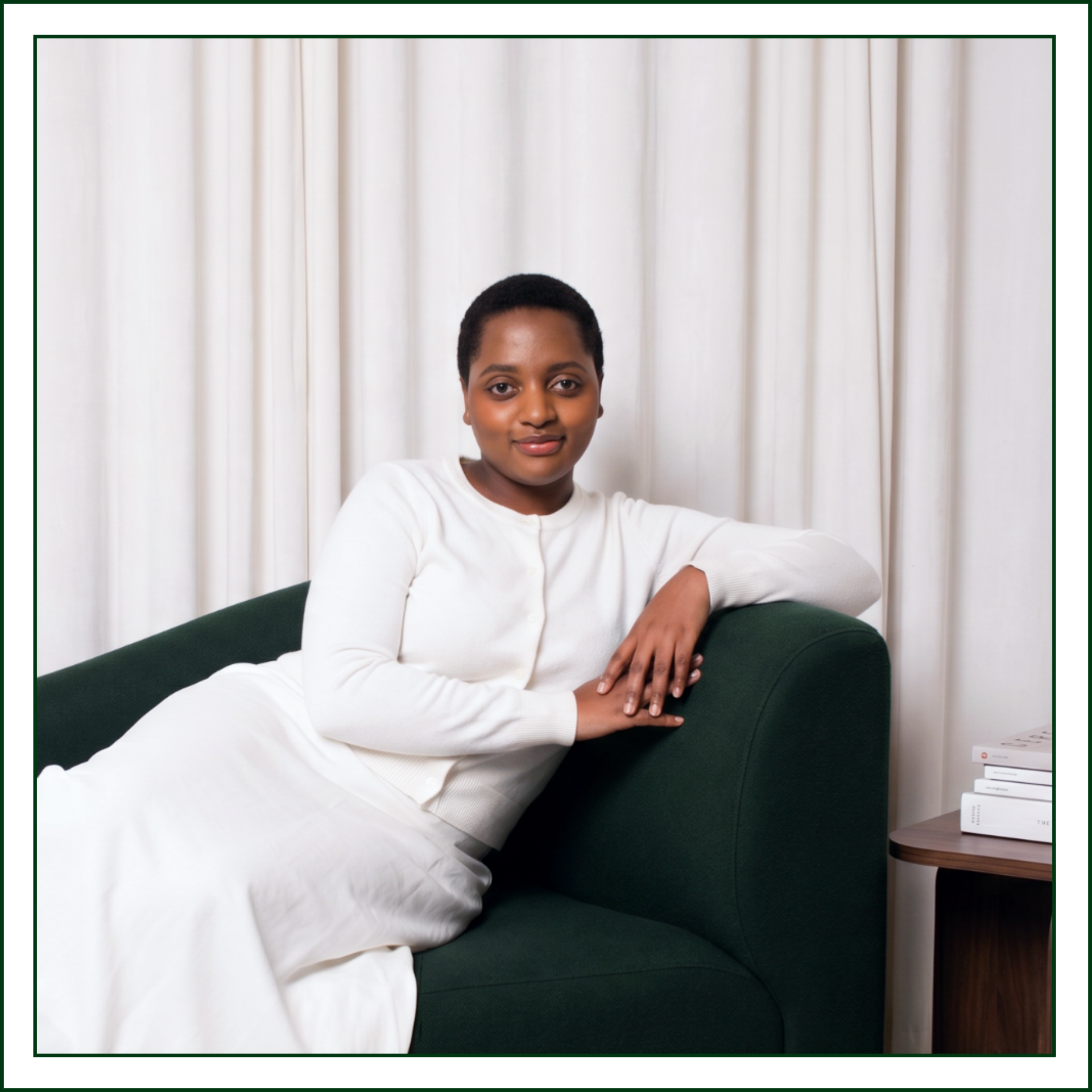 Lillian Ingabire, editorial brand designer and founder of Studio Kalili in Chicago, seated in a green armchair with a stack of design books on a side table beside her.