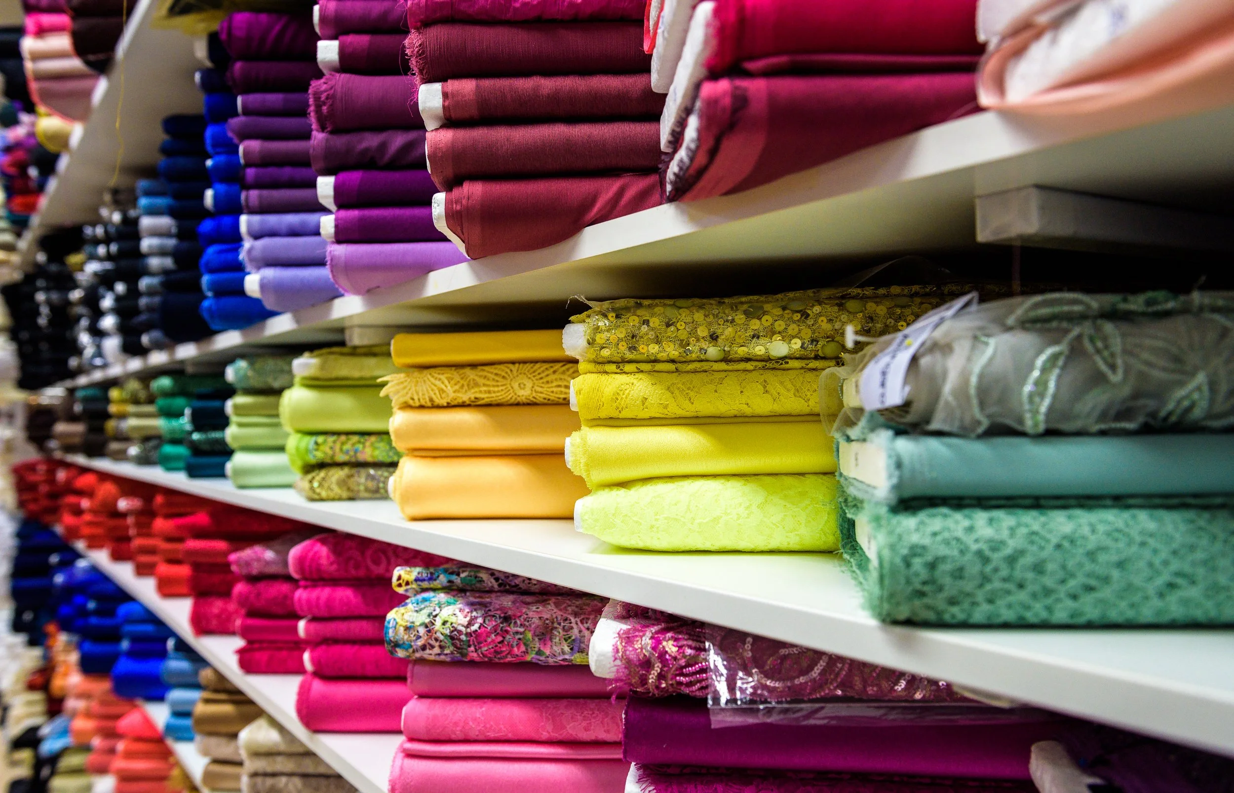 Shelves stocked with various colorful fabrics arranged in rows, including solid colors, floral patterns, and textured fabrics in shades of pink, purple, yellow, green, and blue.
