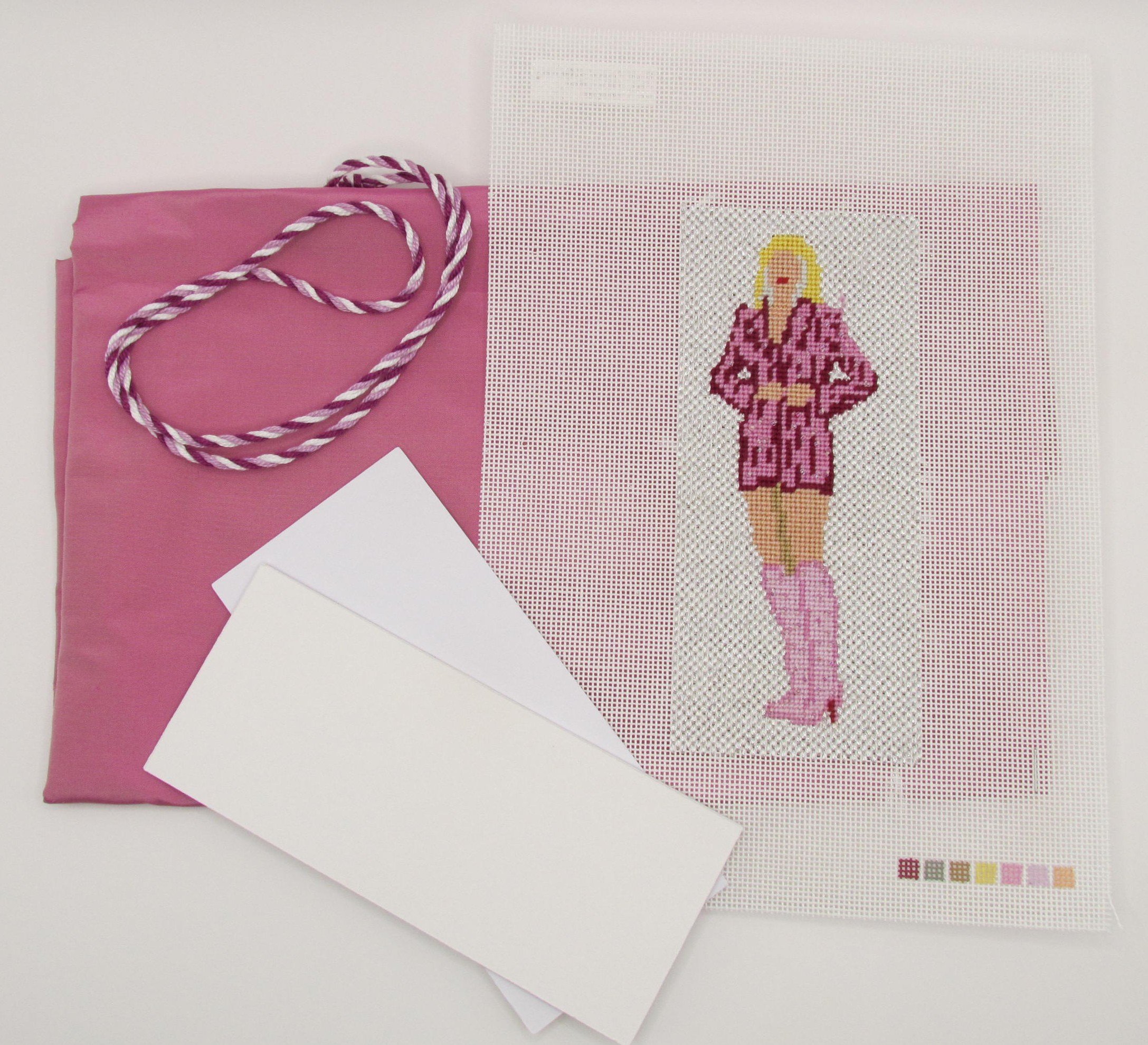 A sewing project featuring a stitched pixel art of a woman with long blonde hair, pink dress, and holding an object, placed on a grid-patterned fabric. The project is accompanied by pink paper, a pink and white striped rope, and white papers surrounding it.