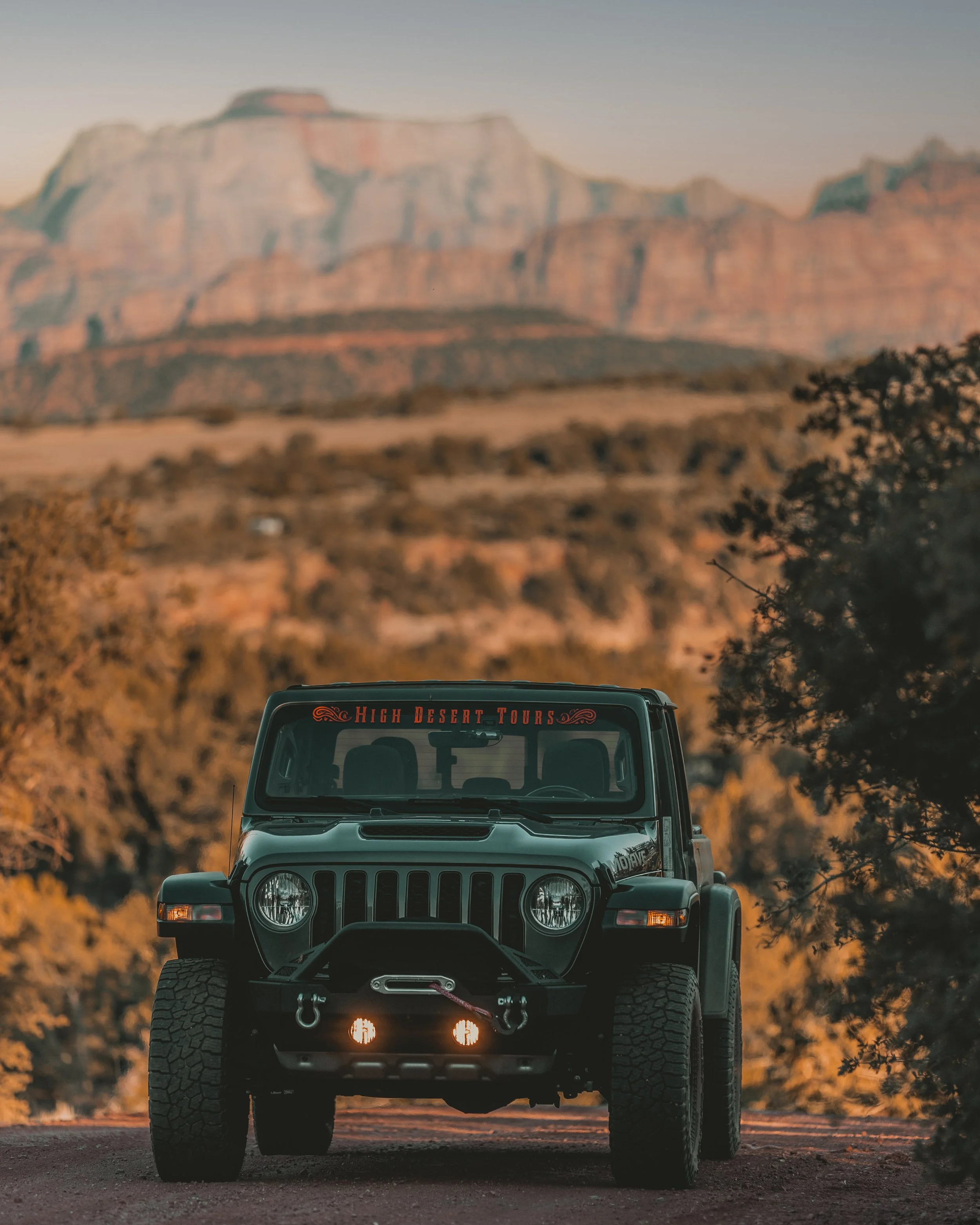 Black Jeep Wrangler with front headlights on, parked outdoors with a mountainous landscape in the background during sunset.