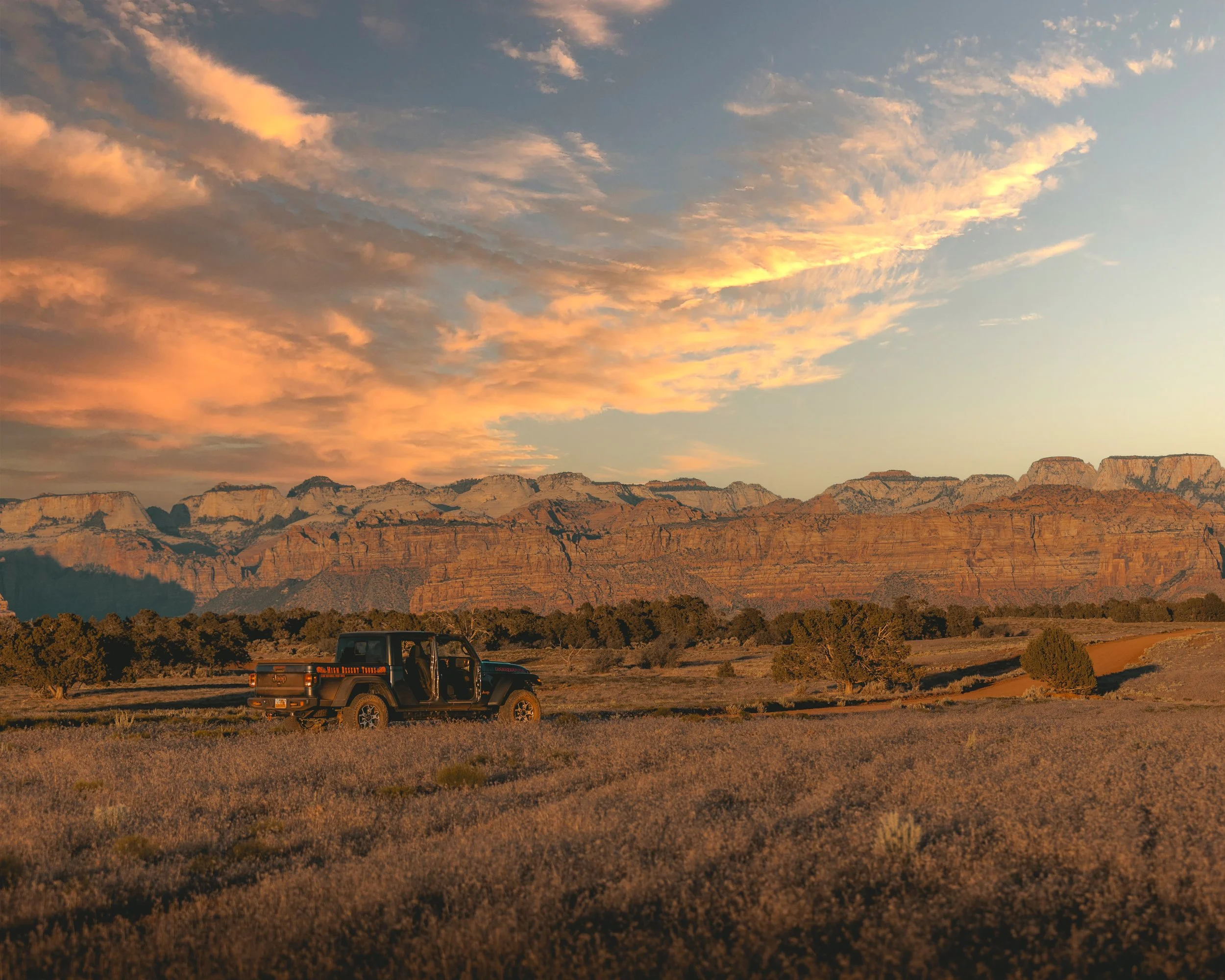 A scenic view of a desert landscape with rocky mesas and cliffs under a partly cloudy sky during sunset. A black Jeep is visible in the foreground on a dirt road.