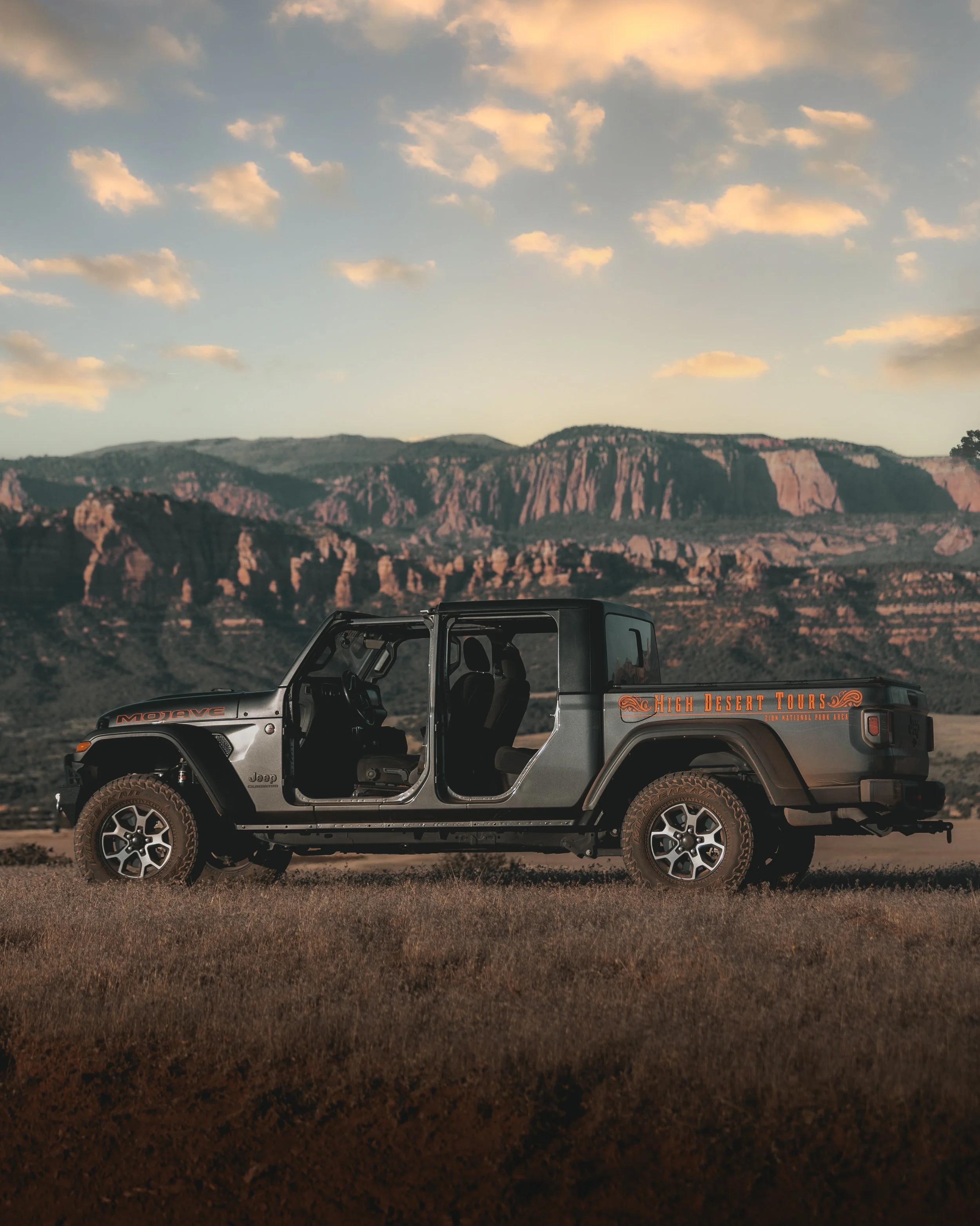 A silver Jeep Wrangler parked on a grassy plain with a mountainous landscape in the background, under a partly cloudy sky at sunset. The vehicle has an orange decal on the side reading 'High Desert Tours'.