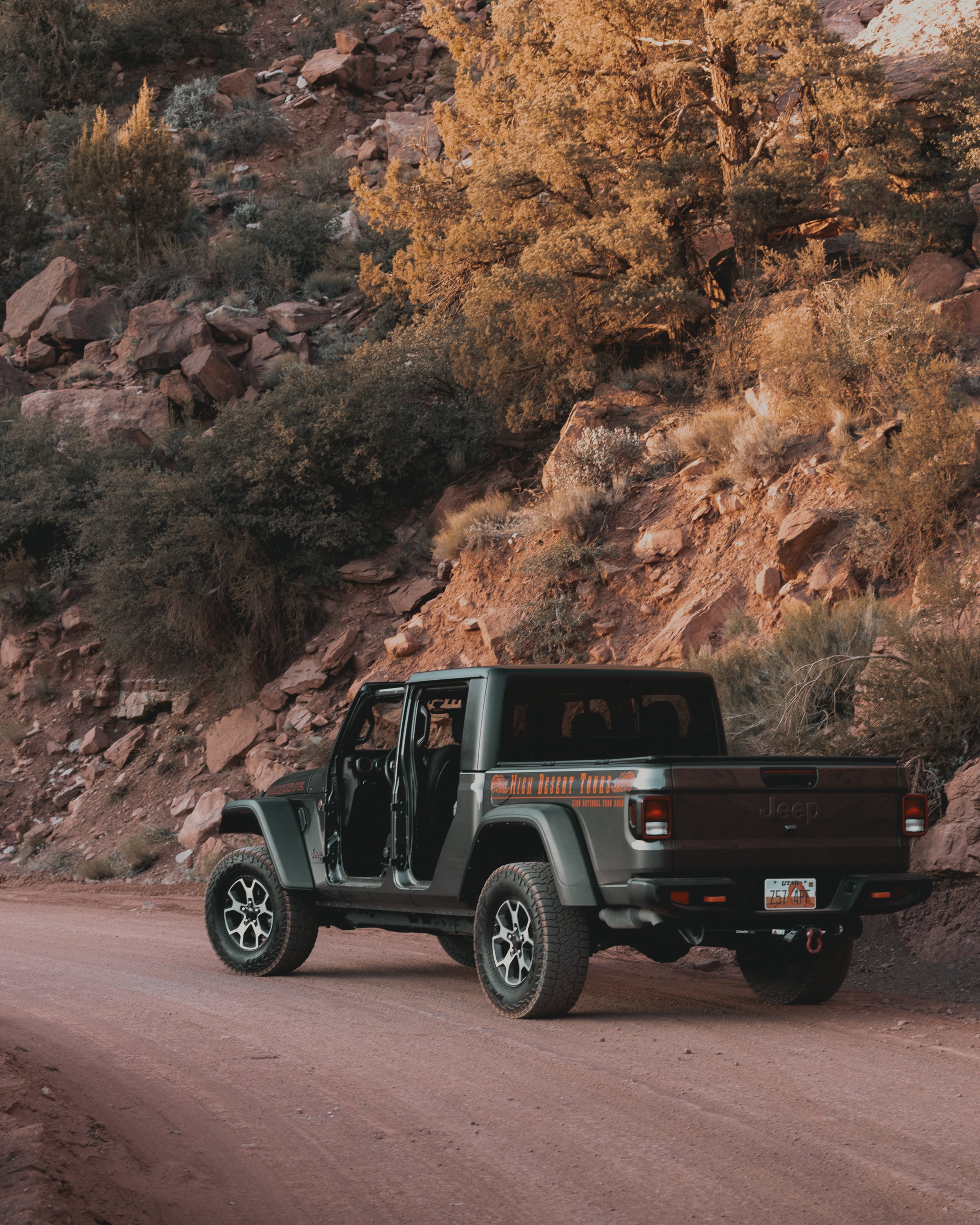 A black Jeep SUV parked on a dirt road in front of a rocky hillside with dry bushes and trees, captured during sunset.
