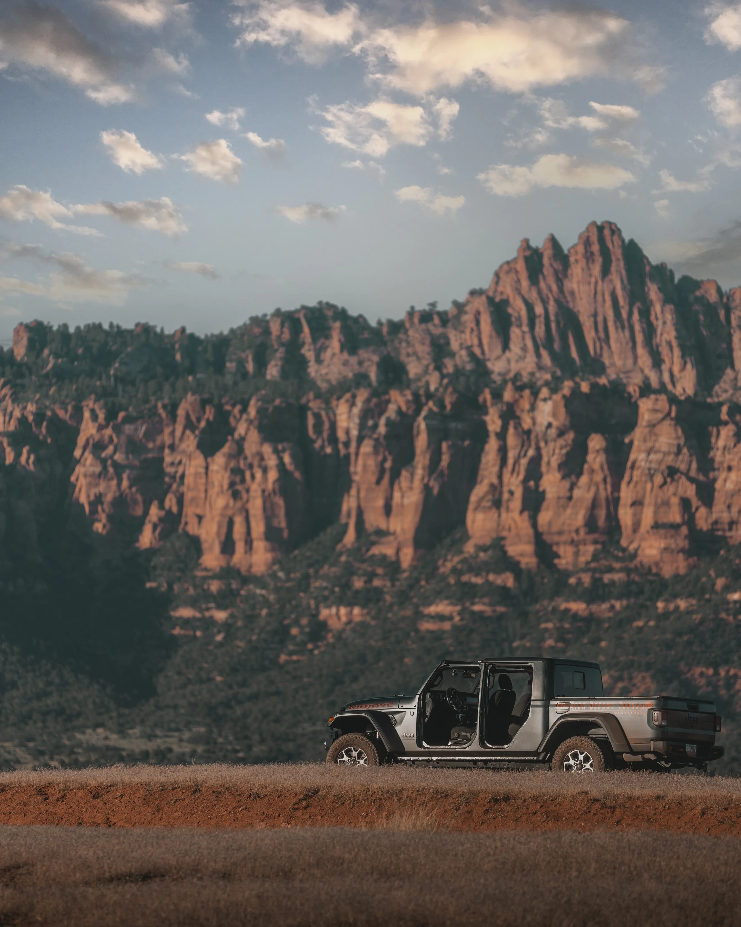 An open black and gray Jeep Gladiator truck parked on a dirt surface in front of a rocky mountain range, with a sky full of scattered clouds overhead.