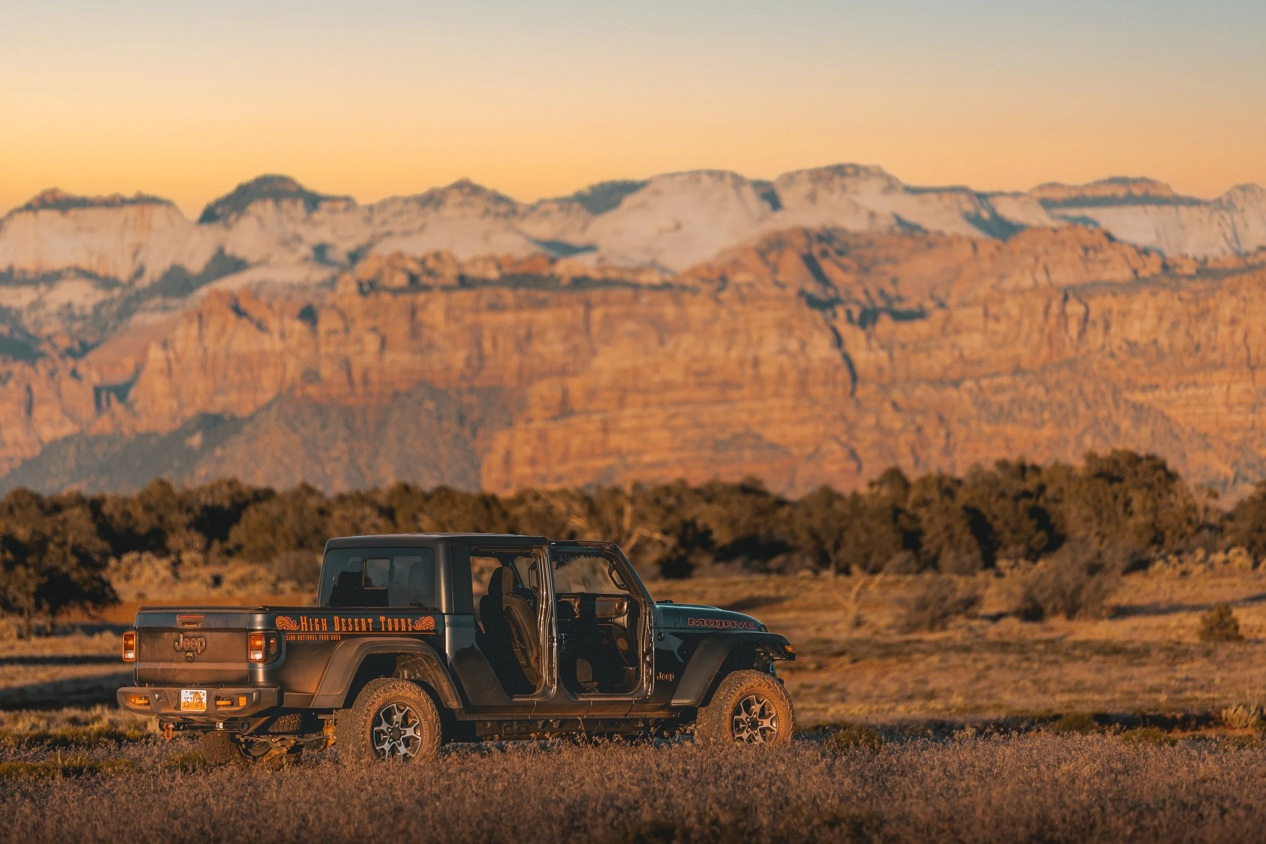 A black Jeep pickup truck parked on a field with mountains in the background during sunset.