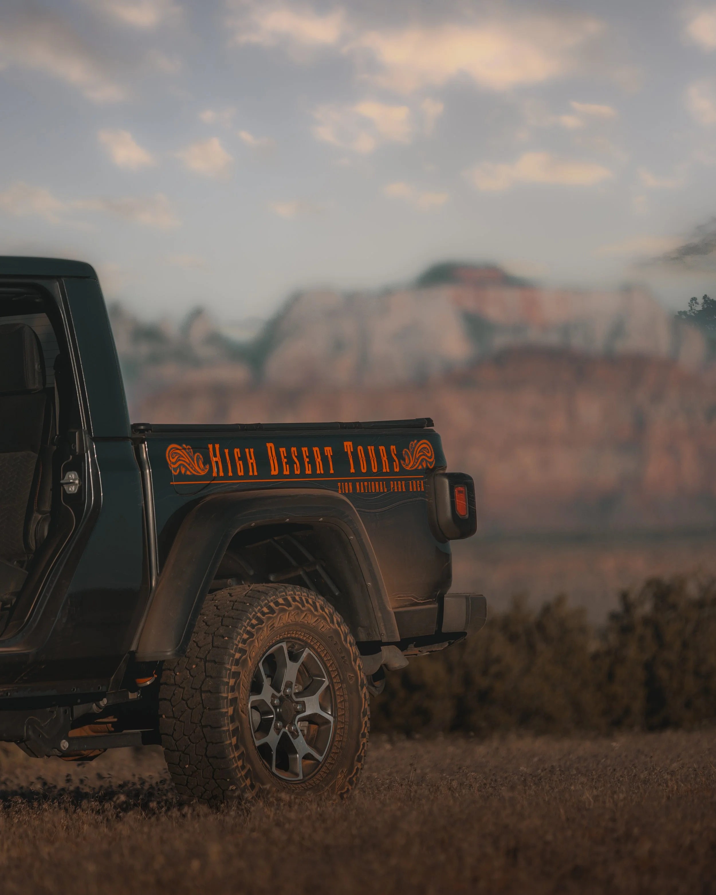 Black off-road vehicle with a sign that reads 'High Desert Tours' on the side, parked on rough terrain with Mount Rushmore in the background under a cloudy sky.